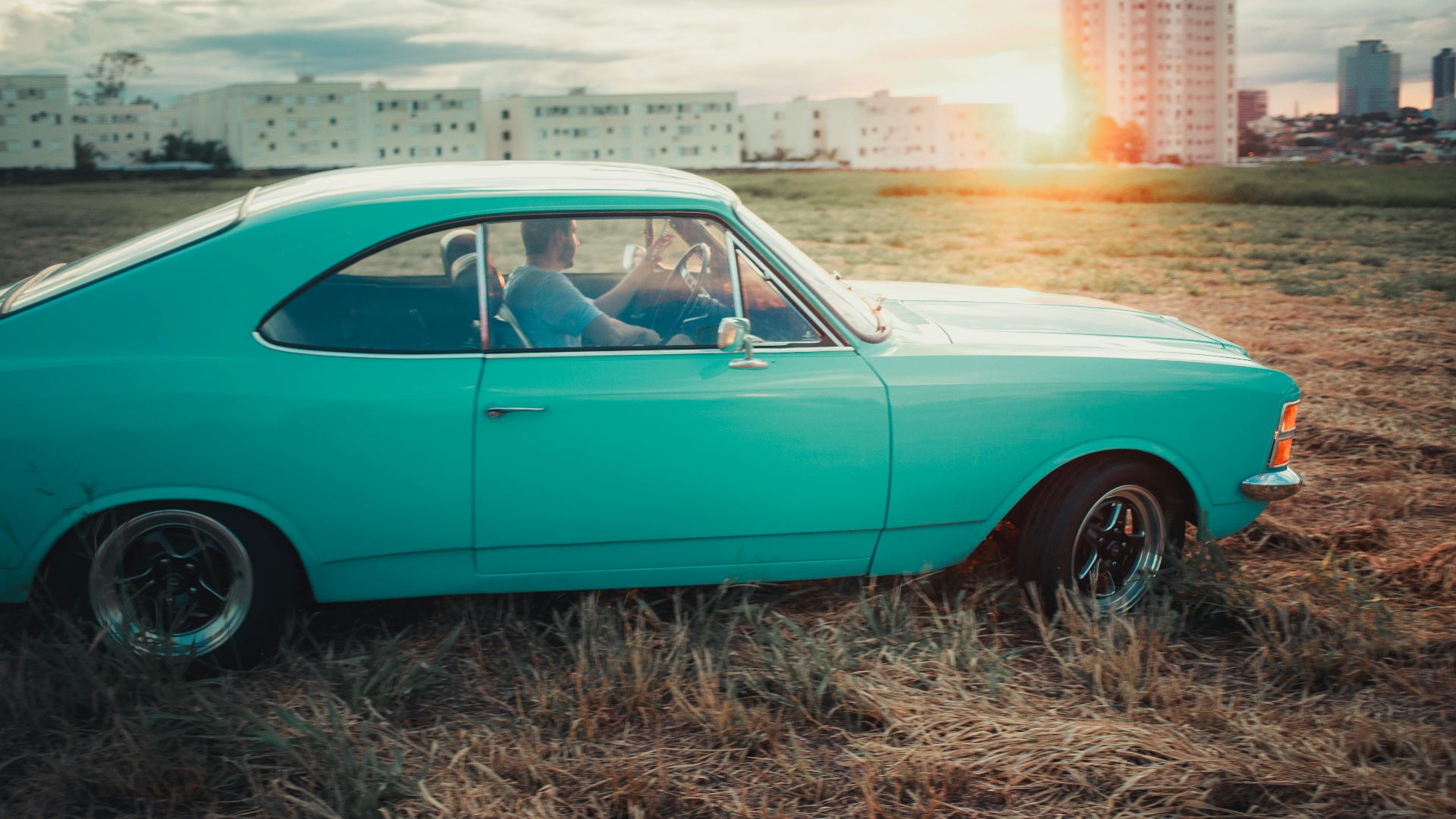 A classic teal car driven by an adult male on a field at sunset, city skyline in background.