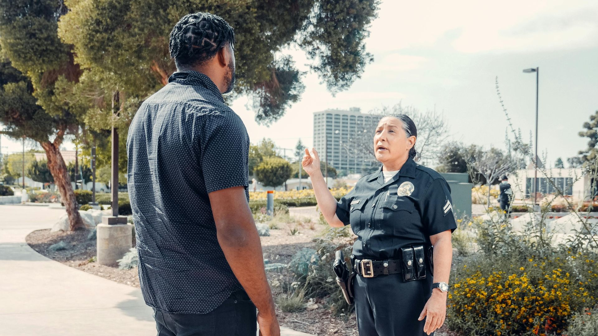 A police officer engages in conversation with a civilian in a park setting on a sunny day.