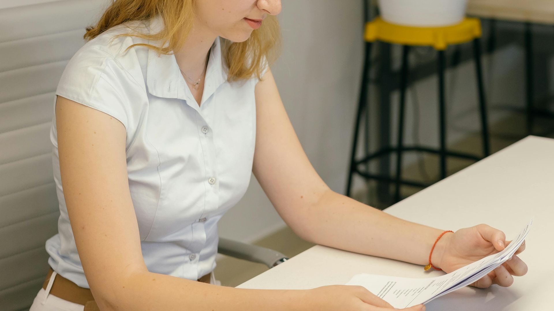 A woman in a modern office reading documents at a desk beside an indoor plant.