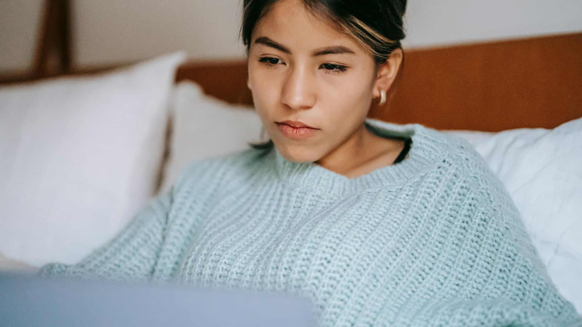 Crop focused ethnic female in warm clothes with portable computer resting on bed at home