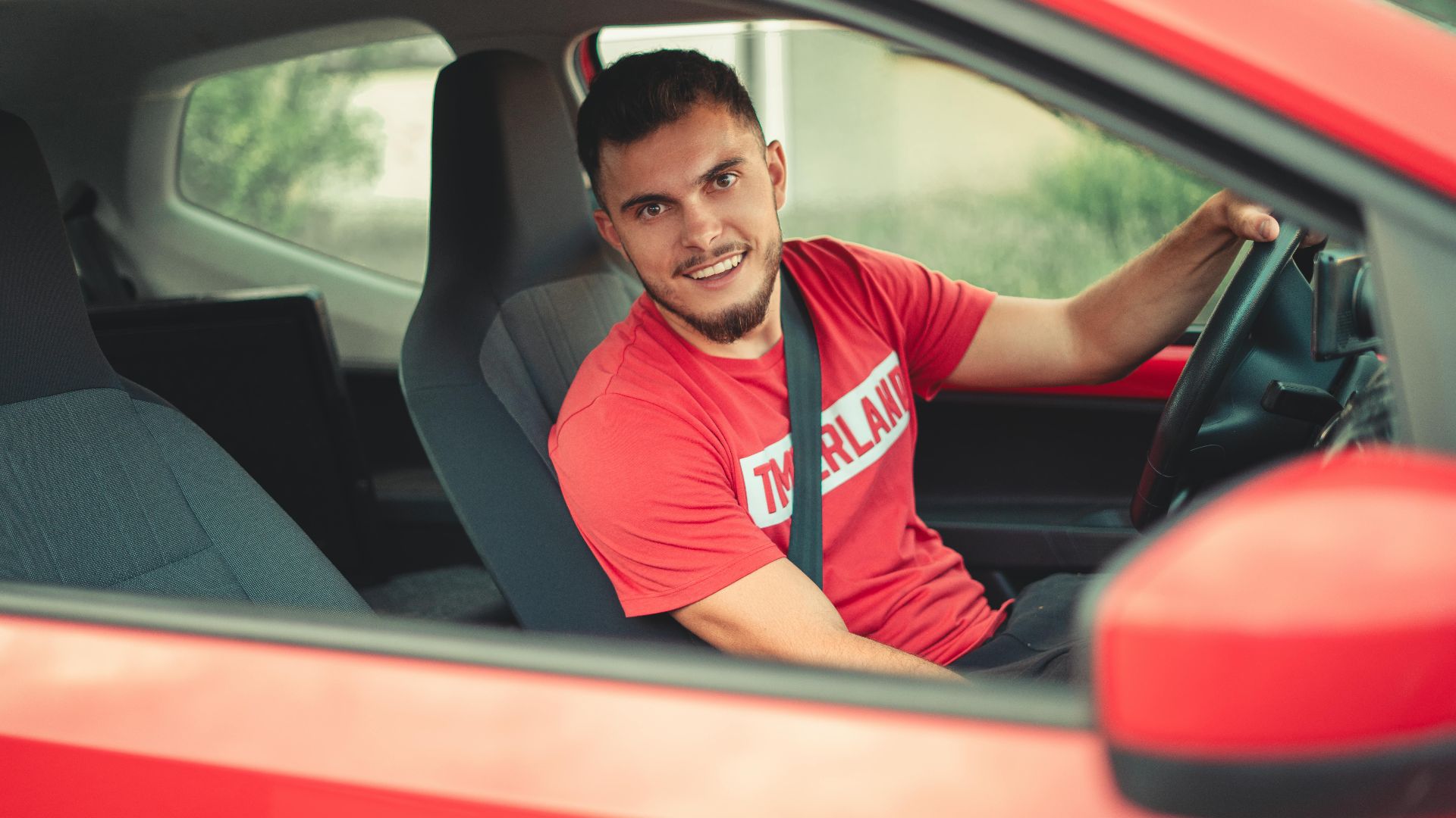 Smiling man driving a red car while wearing a seatbelt, showcasing safety and joy.