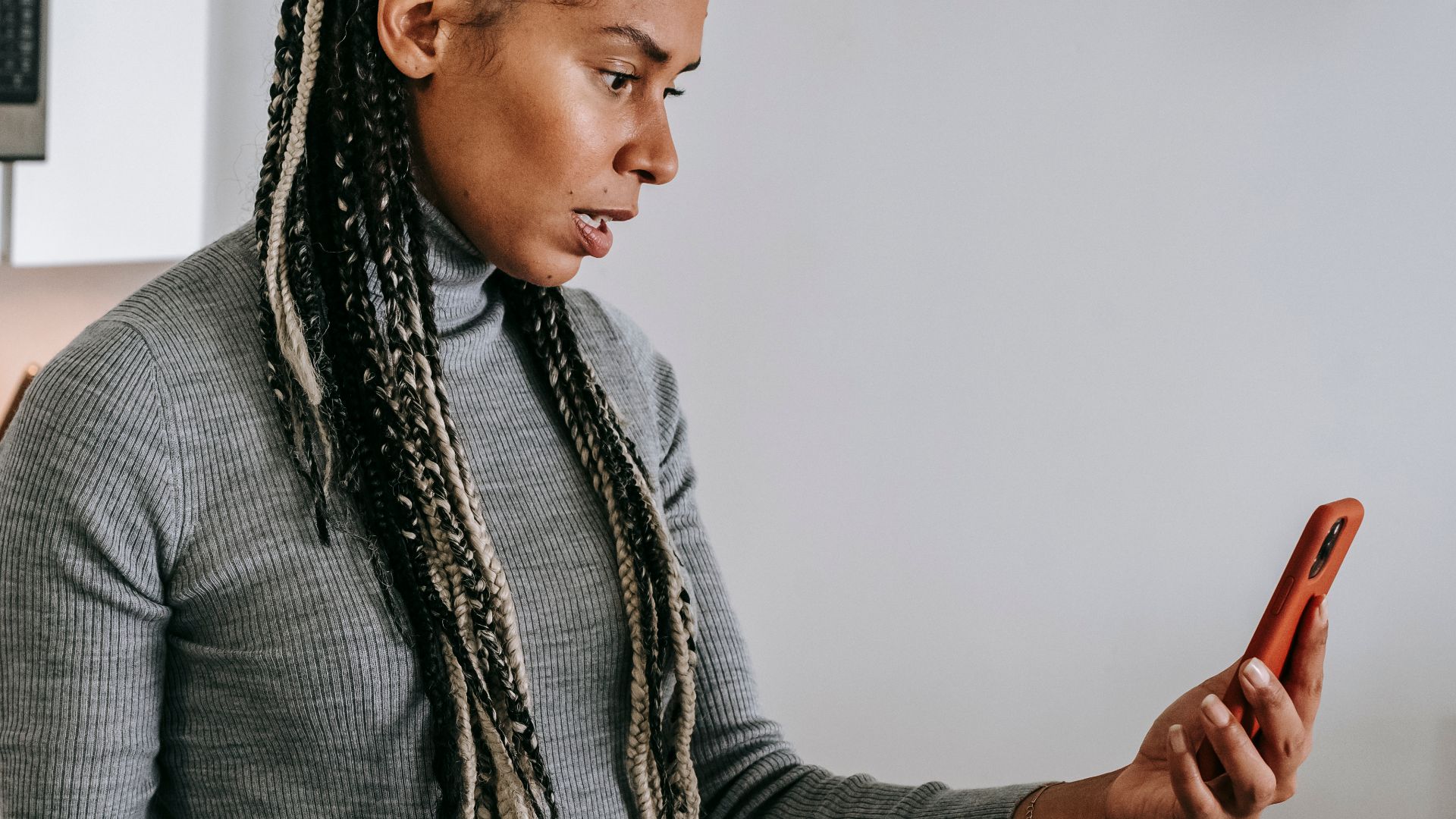 Serious young black lady with Afro braids in casual clothes gesticulating while having unpleasant conversation via video chat on smartphone in modern kitchen
