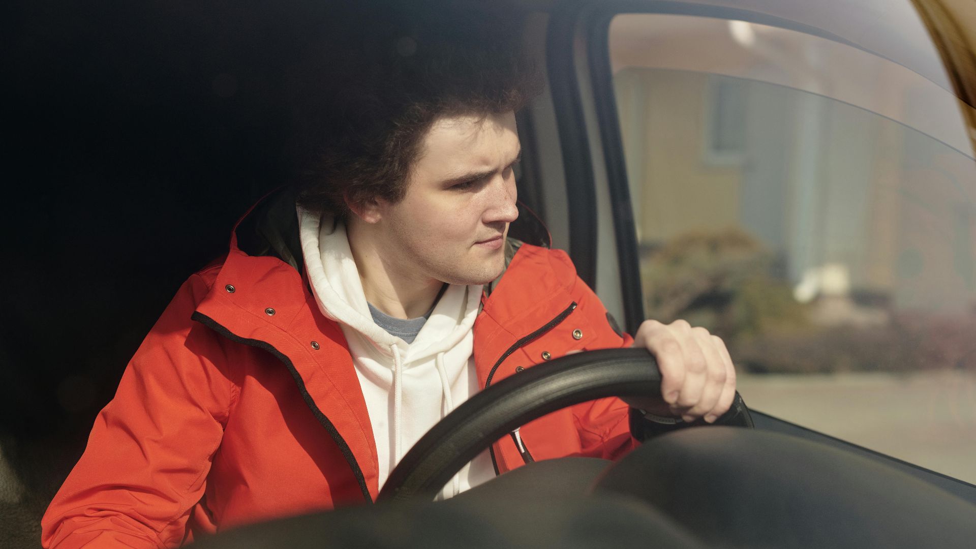 A young man in a red jacket steering a car with focus, enjoying a peaceful drive.