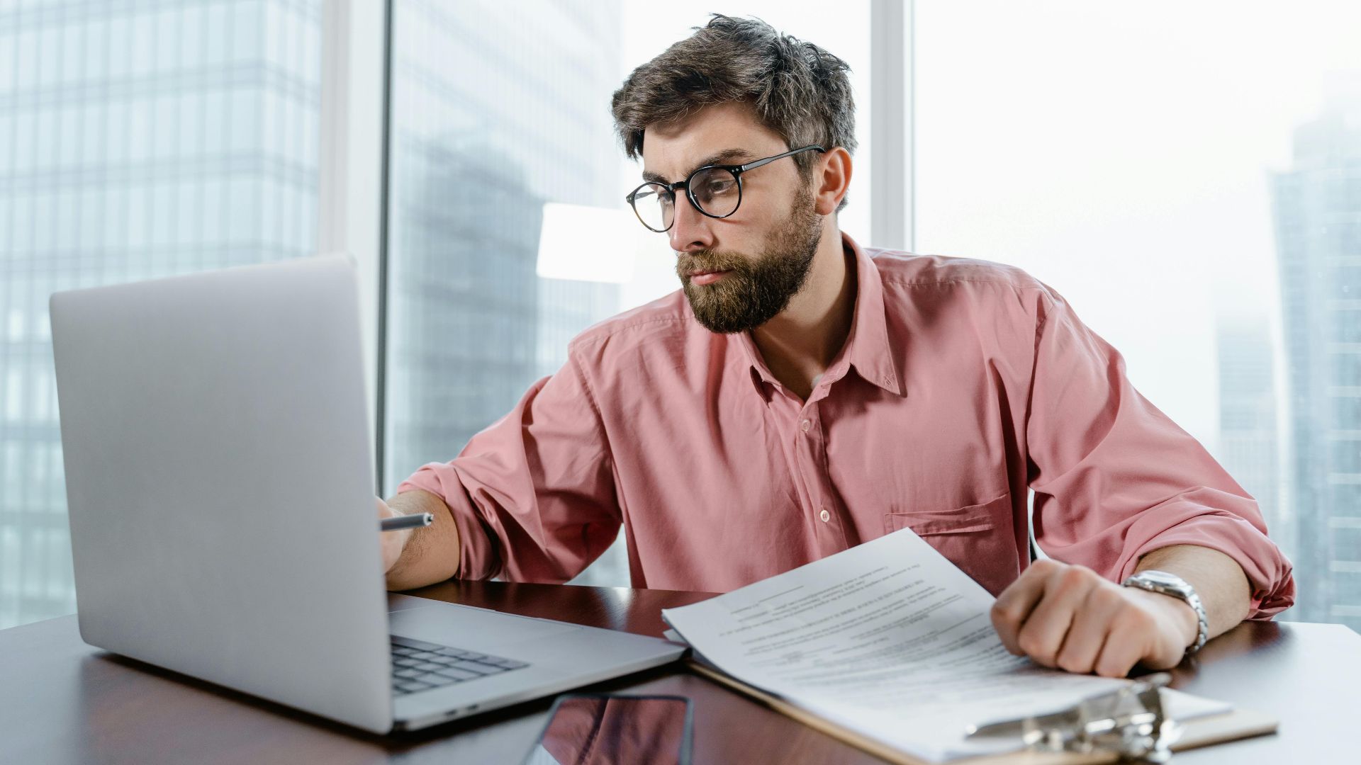 A businessman in an office reviews documents and data on a laptop, concentrating on financial analysis.
