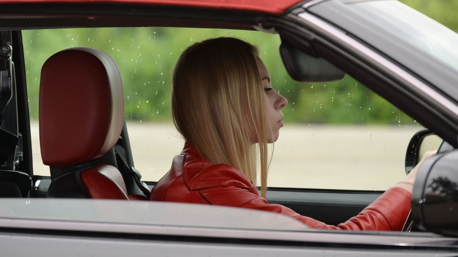 A young woman in a red convertible driving on a rainy day, wearing a red jacket.