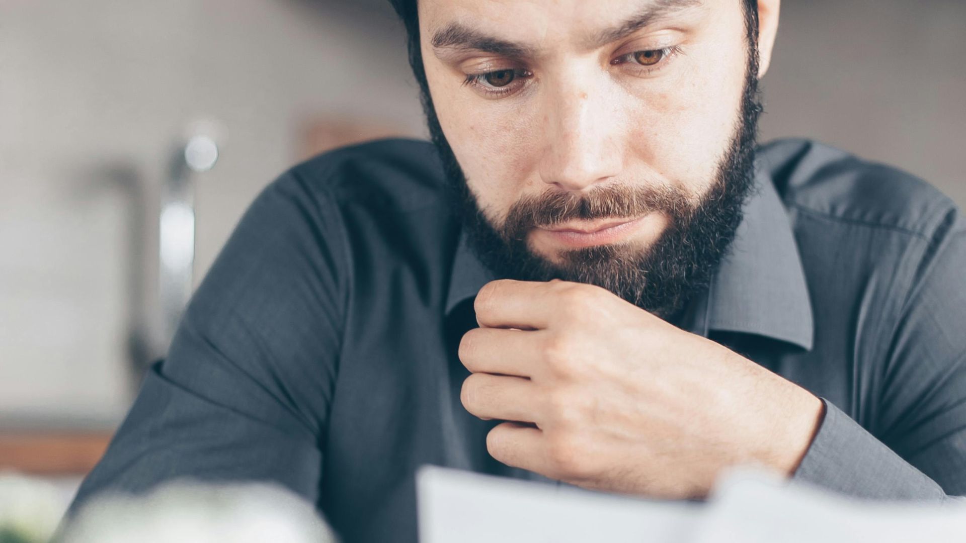 A bearded man in a gray shirt intently reads documents while seated indoors, appearing concerned.