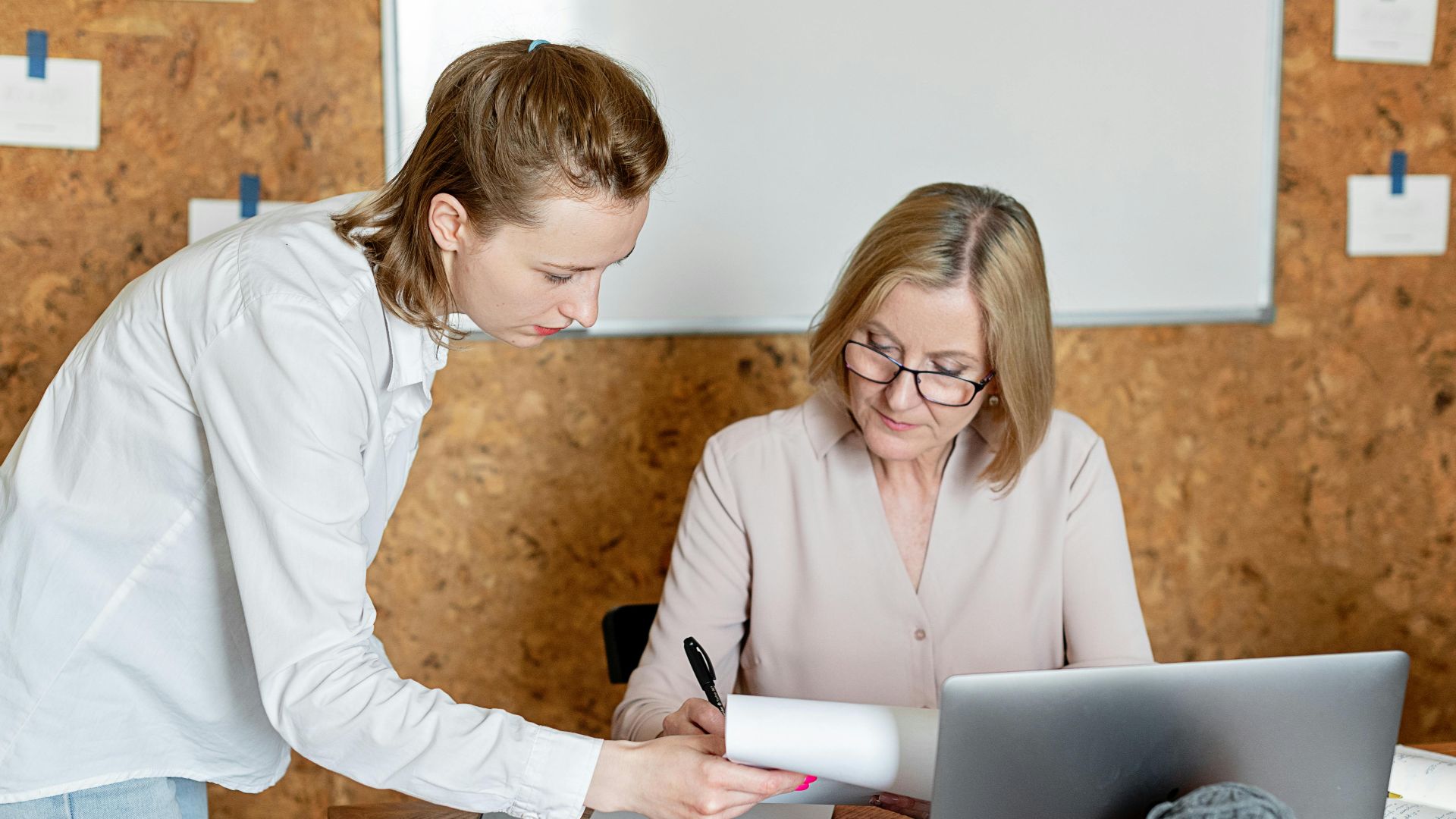 Two women discussing work documents in a modern office setting with a laptop.