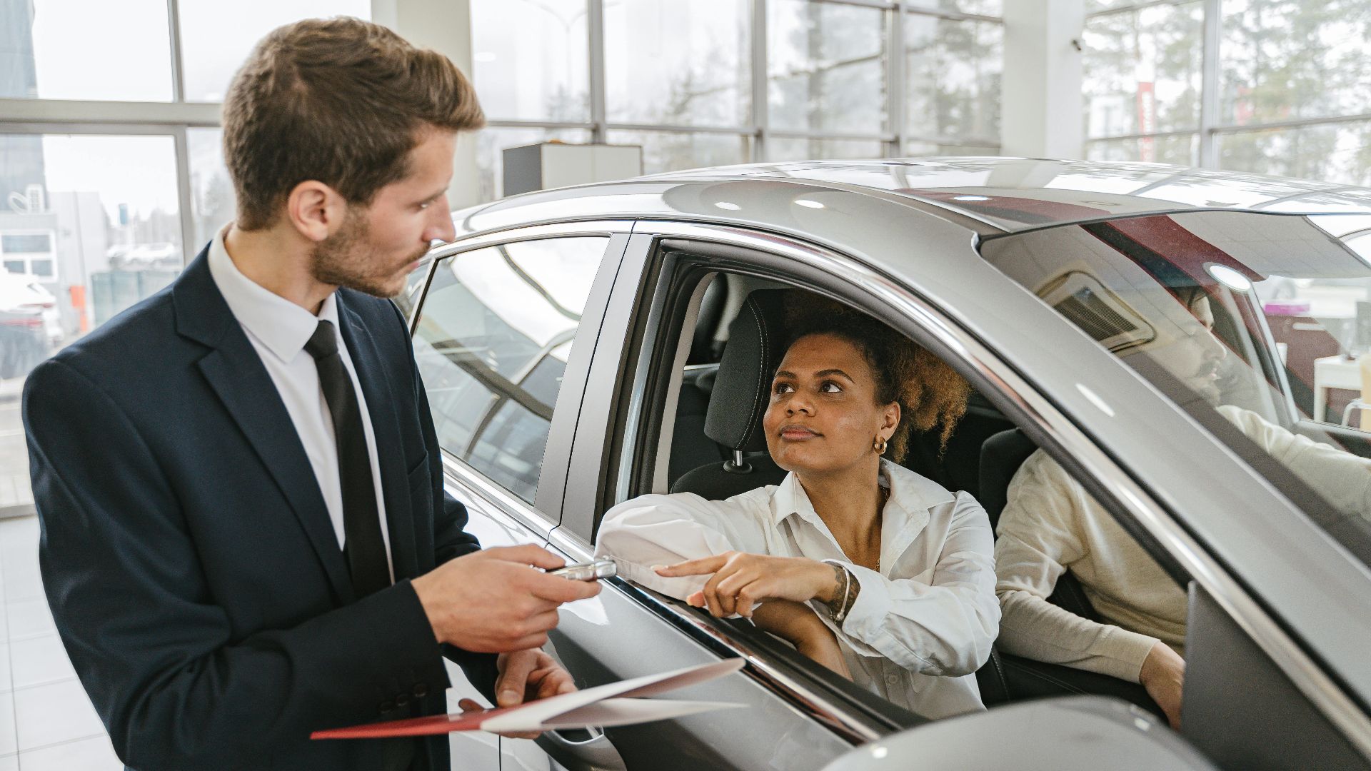 A car dealer hands keys to a woman sitting inside a vehicle in a car showroom.