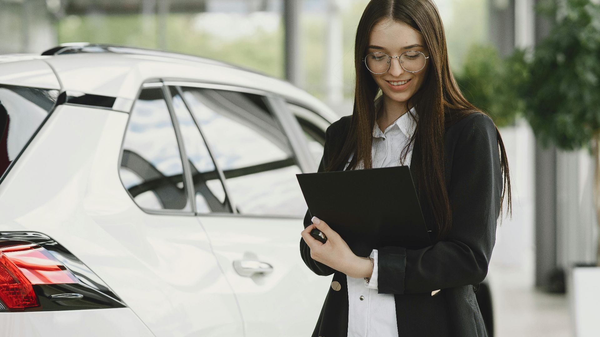 Smiling woman in a black jacket reviewing documents near a white car indoors.