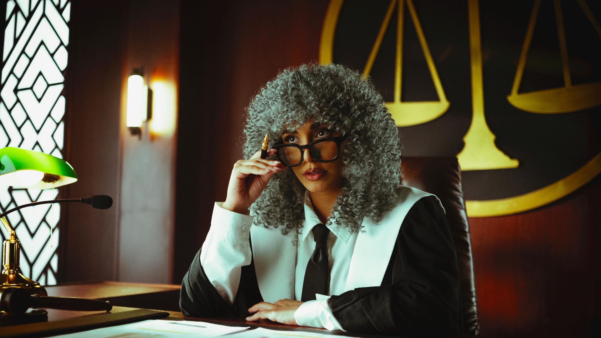 Judge in traditional attire sits at a courtroom desk with a serious expression.