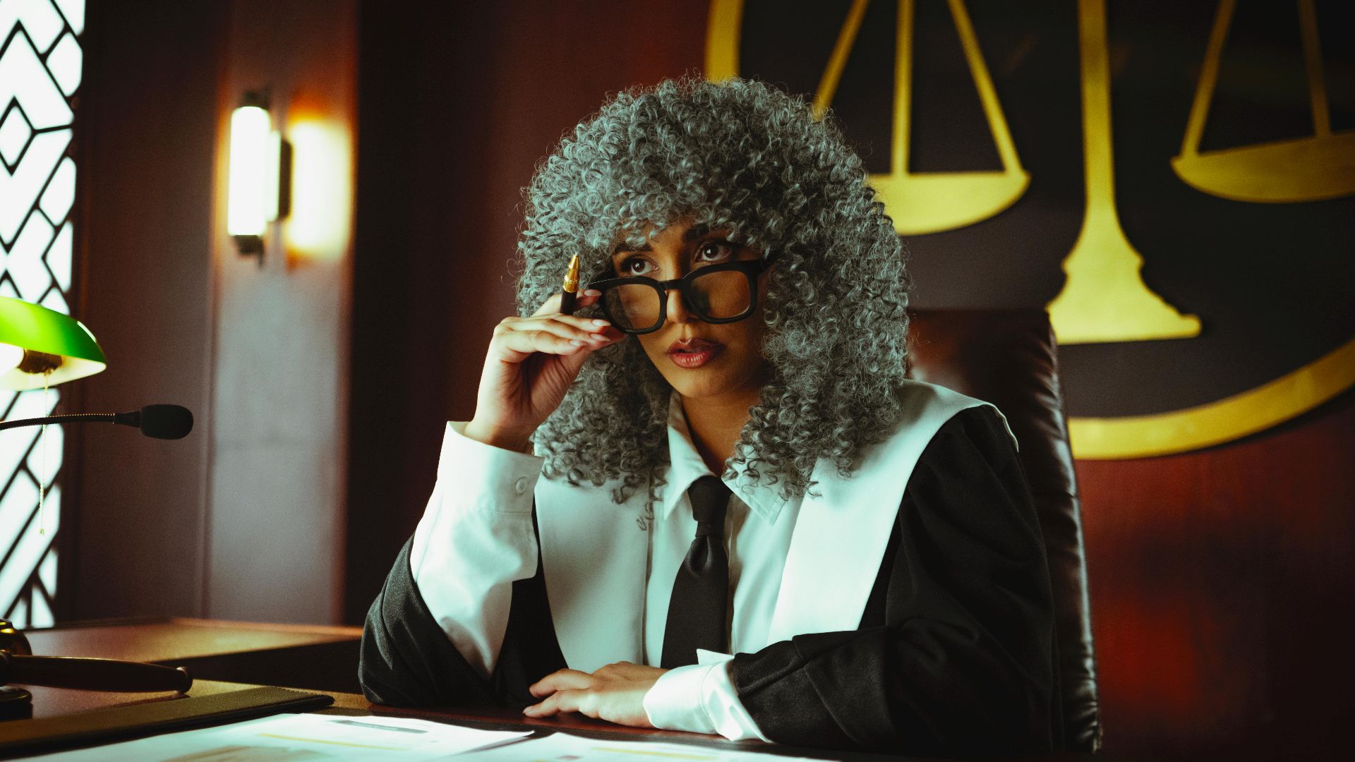 Judge in traditional attire sits at a courtroom desk with a serious expression.