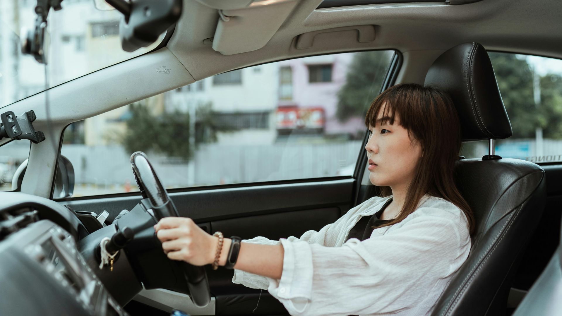 Side view unemotional Asian female in casual white clothes steering right hand drive car along urban streets in daytime
