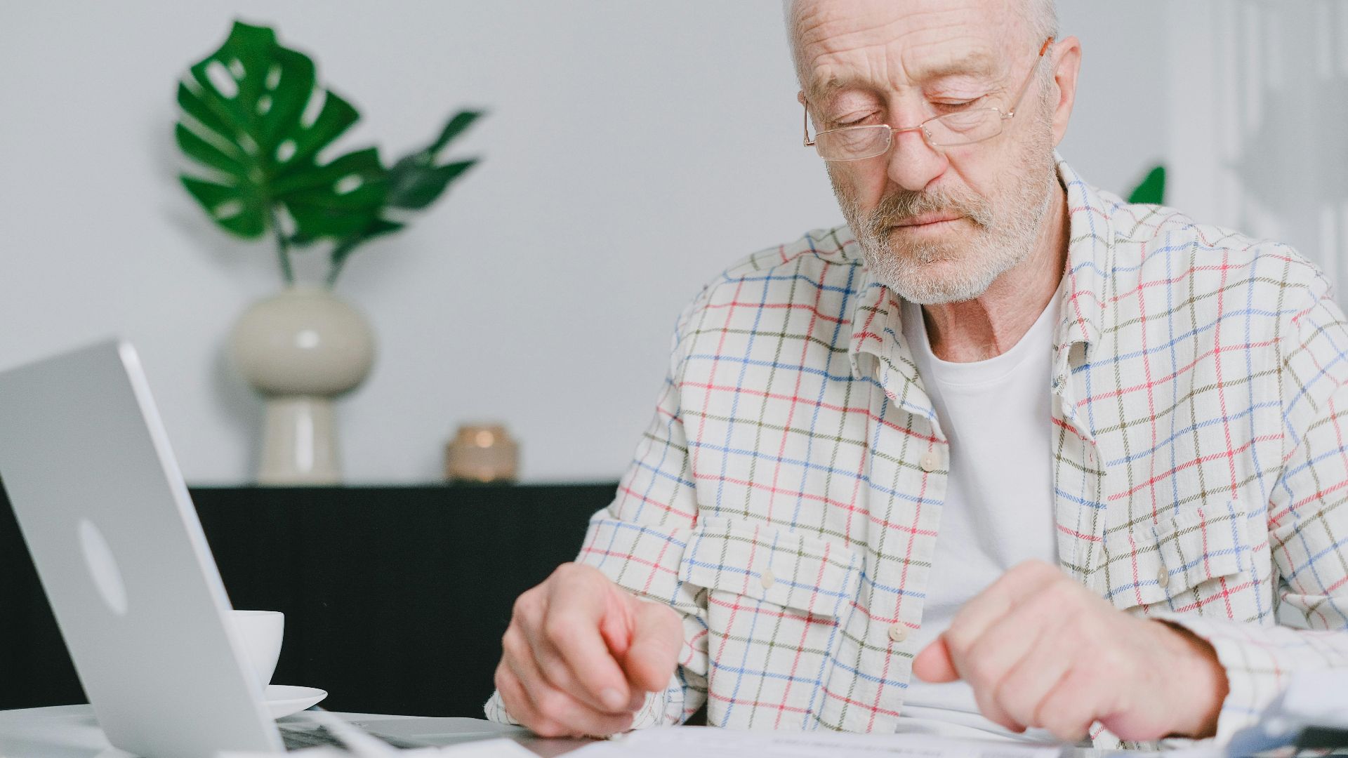 Elderly man with eyeglasses focused on paperwork at home, using laptop.