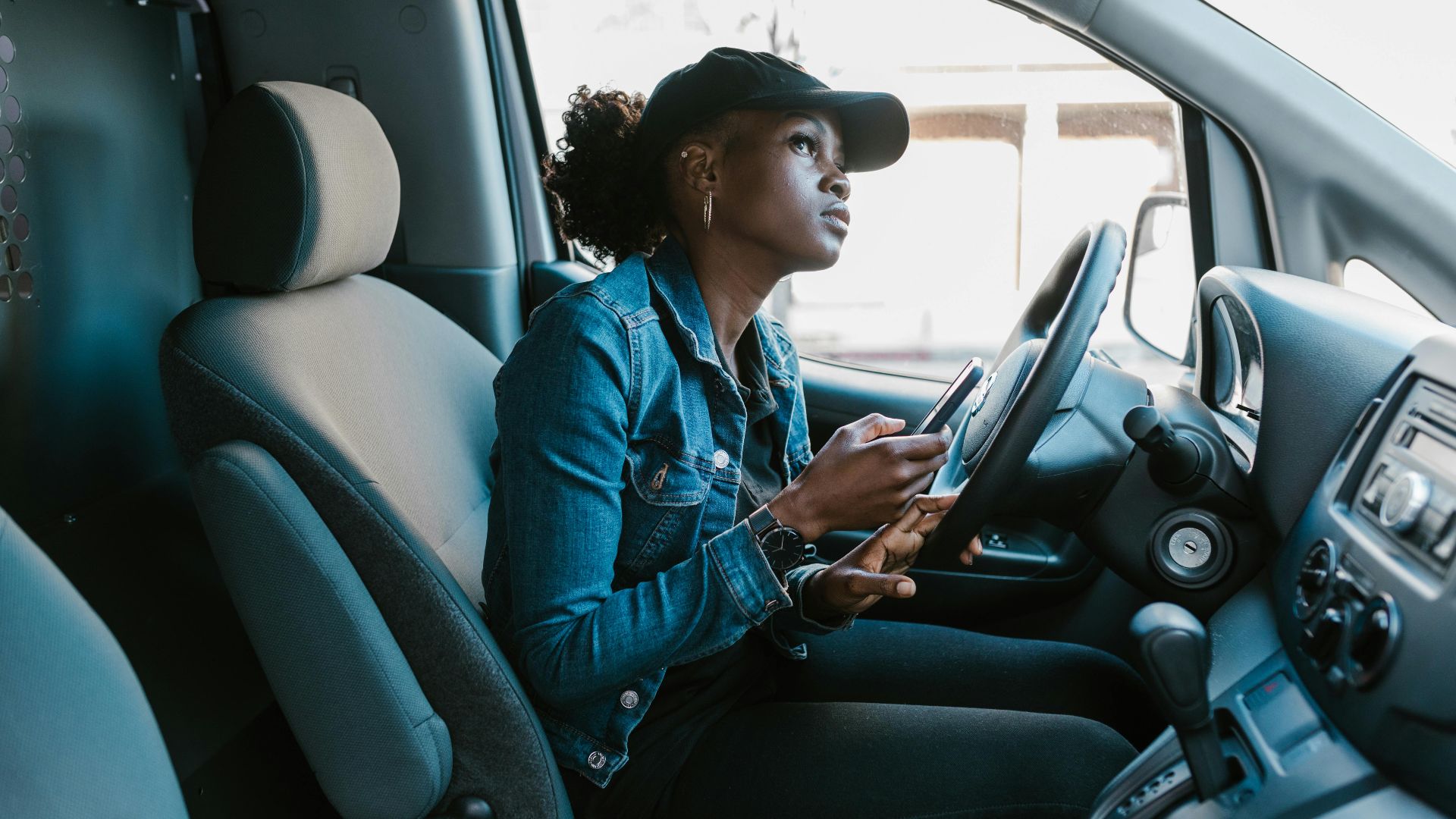 A woman in a denim jacket focuses on her smartphone while sitting in a car driver's seat.