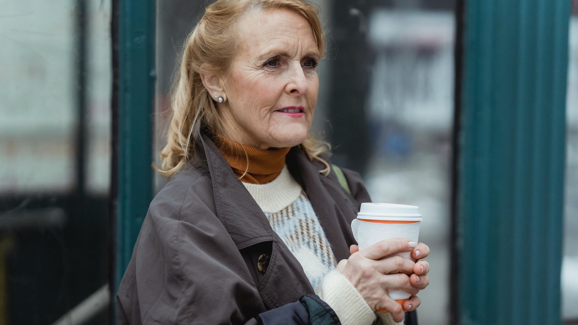 Contemplative elderly female in outerwear with hot beverage to go looking forward in town in daylight