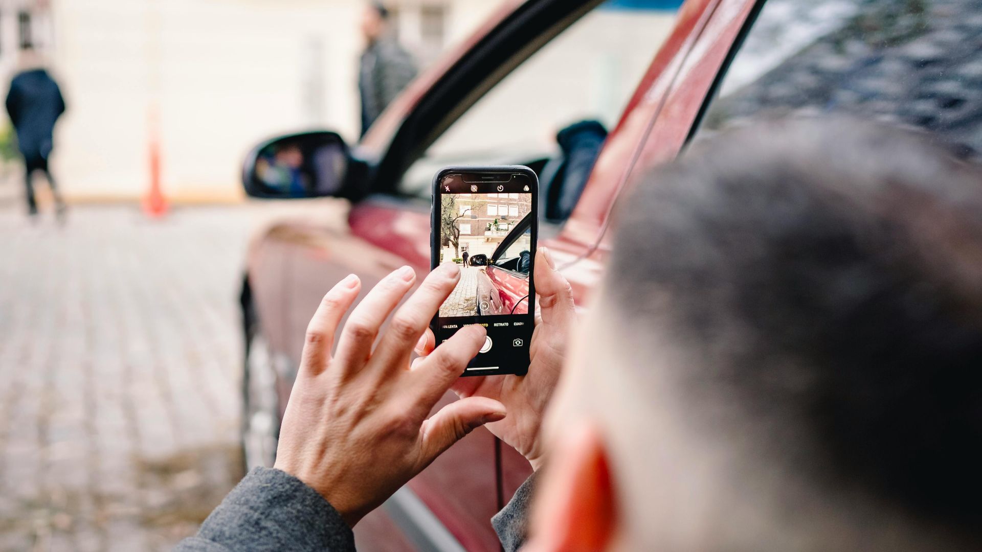 Man using smartphone to take pictures of an urban scene beside a red car.
