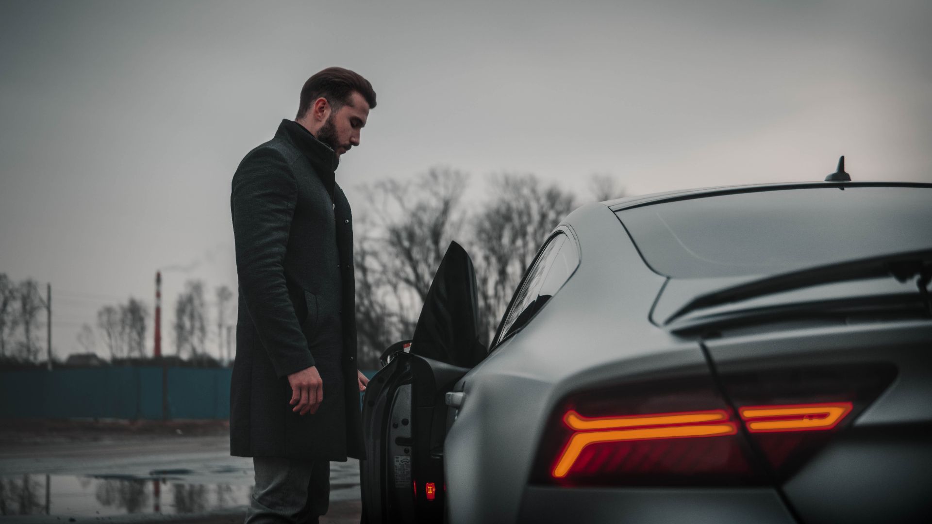 A man in a black coat stands beside a luxury car on a cloudy day in Moscow, Russia.