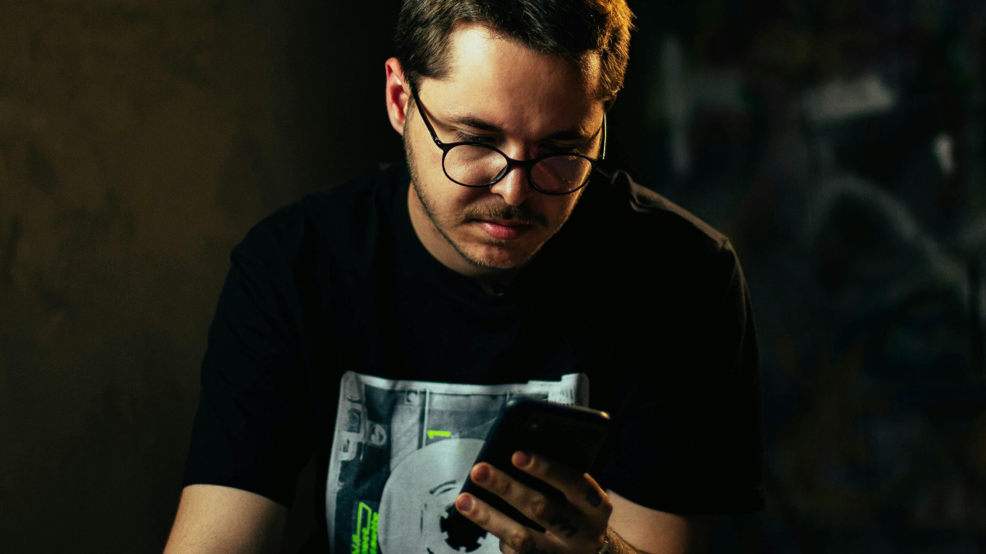 Young man with eyeglasses focused on smartphone indoors in moody lighting.