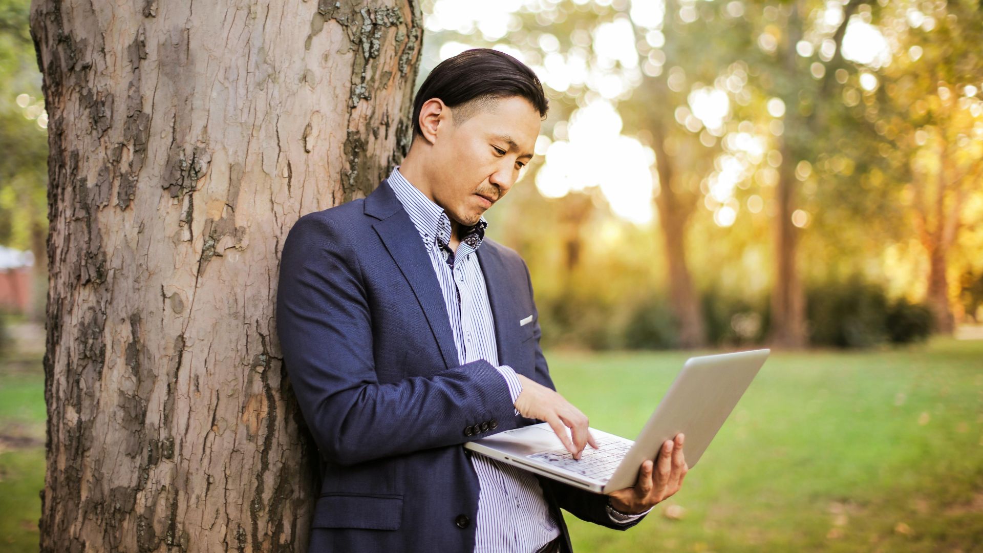 Asian man using a laptop in a park, symbolizing remote work and digital nomad lifestyle.