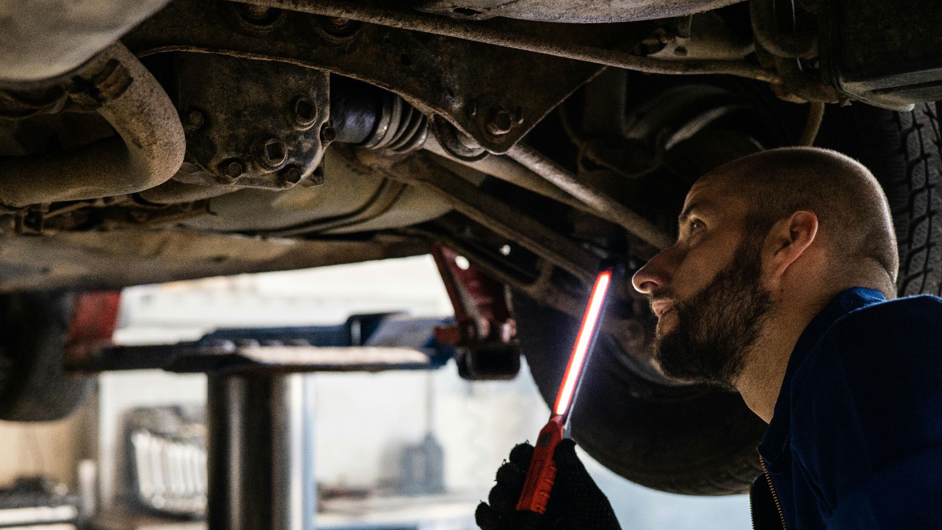 Mechanic using light to inspect vehicle undercarriage in workshop for maintenance.