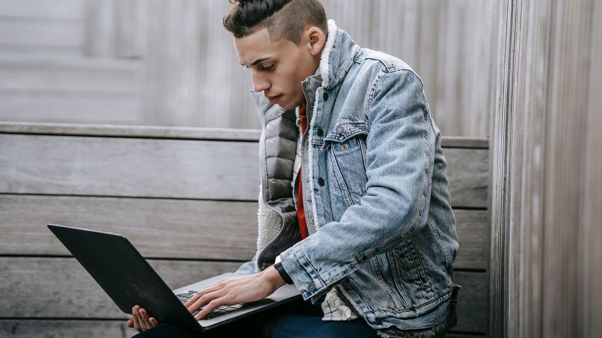 Side view of young male with unusual haircut in denim fashionable jacket browsing netbook on street