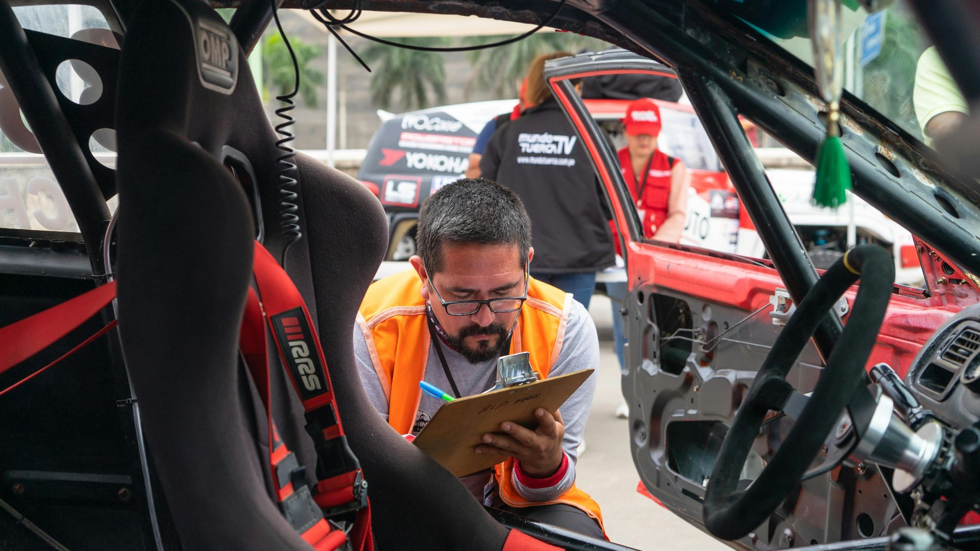 A man inspects a race car interior at a motorsport event, noting findings on a clipboard.