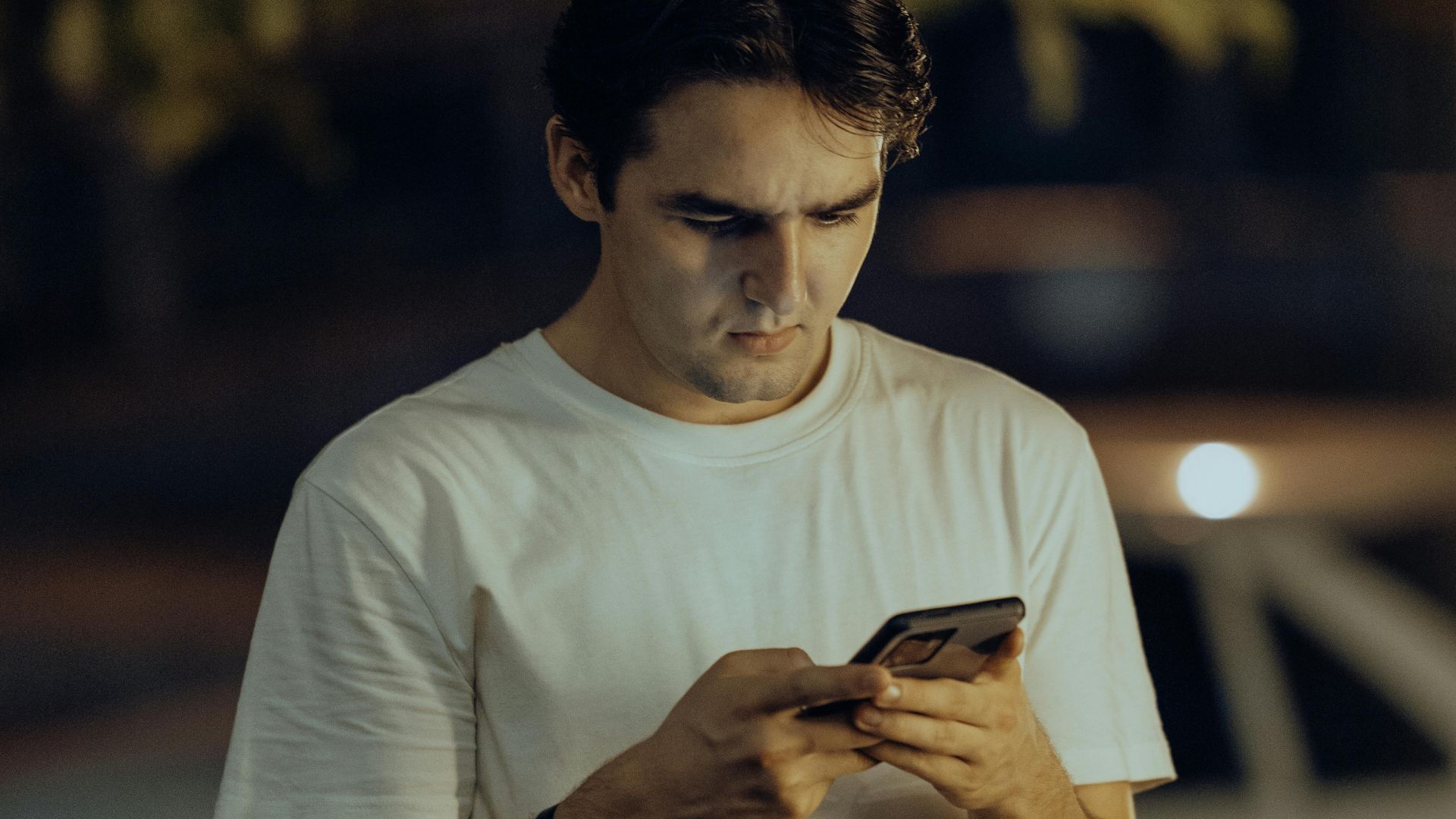 A young man in a white shirt stands outdoors using his smartphone during nighttime.
