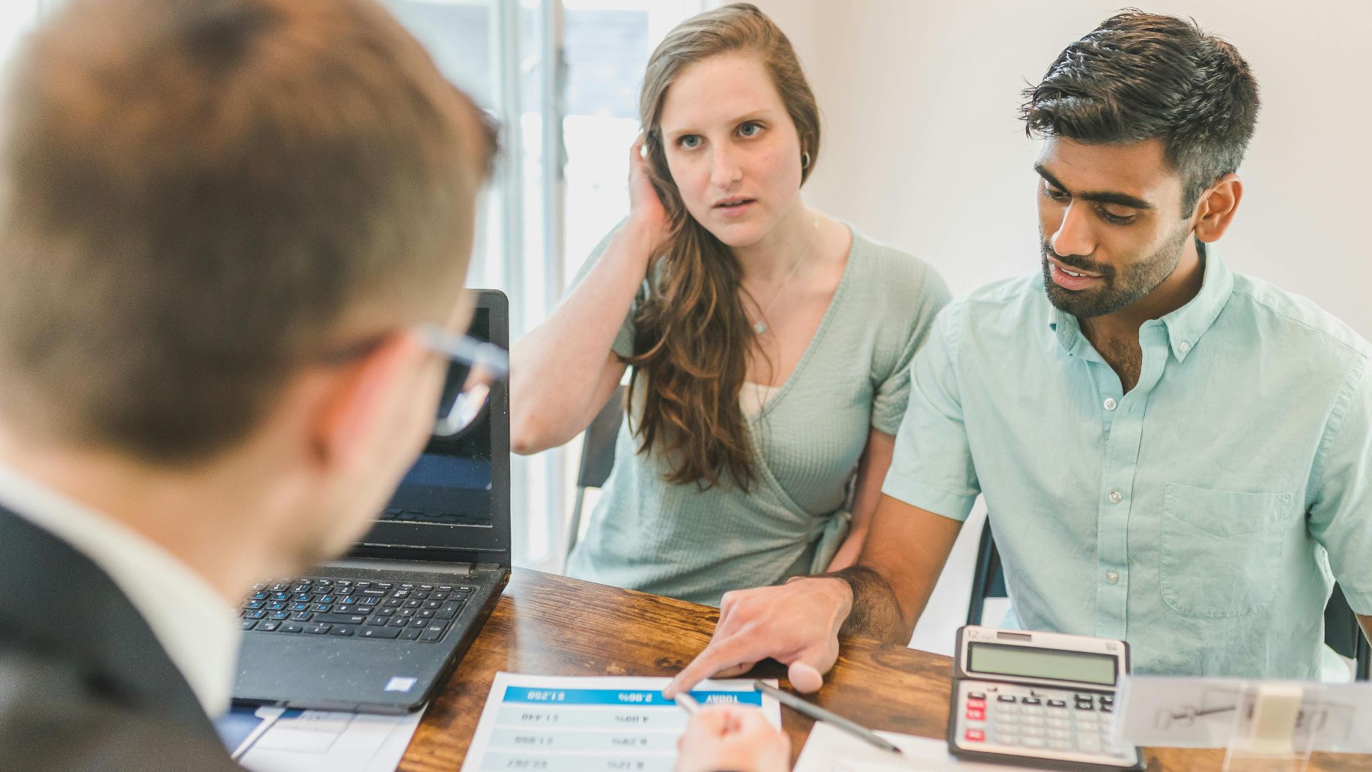 A young couple consults with a realtor about buying a home, indoors in a modern office setting.