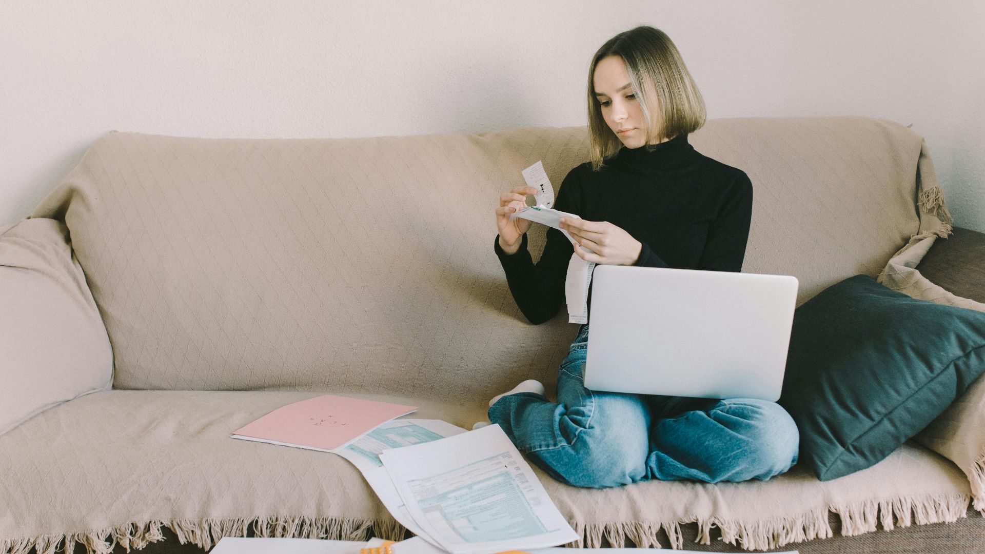 A woman working from home on a sofa with a laptop and documents, managing finances.