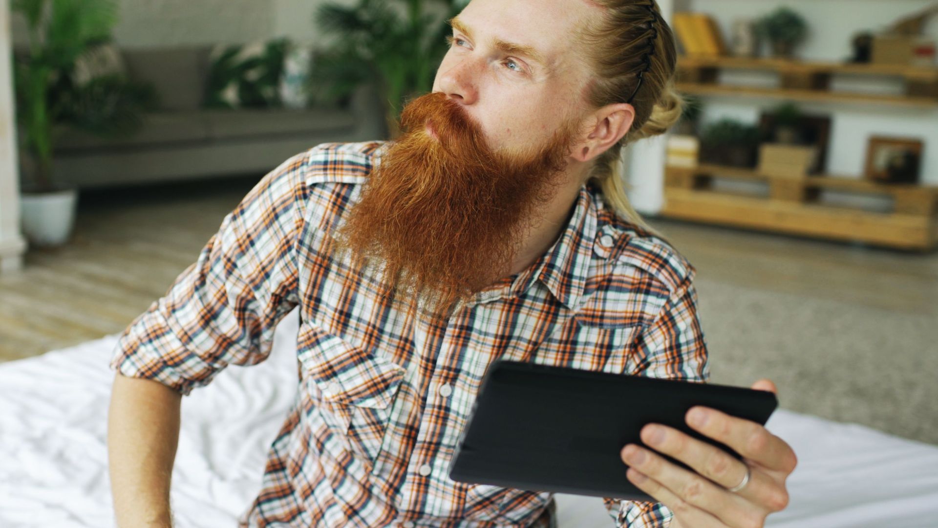 Pensive man with a red beard holding a digital tablet, sitting in a cozy indoor setting.