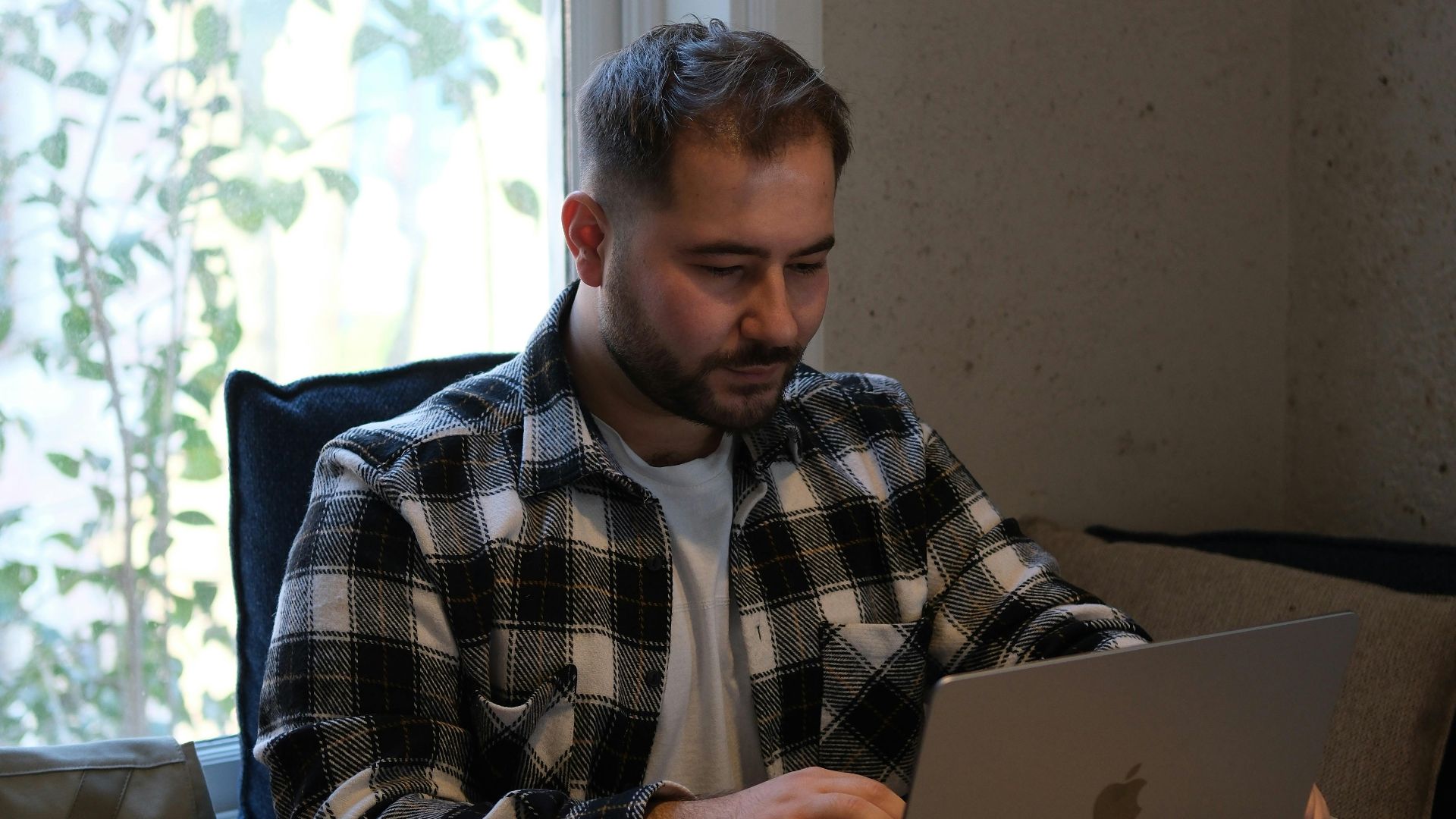 Adult man in checkered shirt working on laptop at a desk in a contemporary office environment.