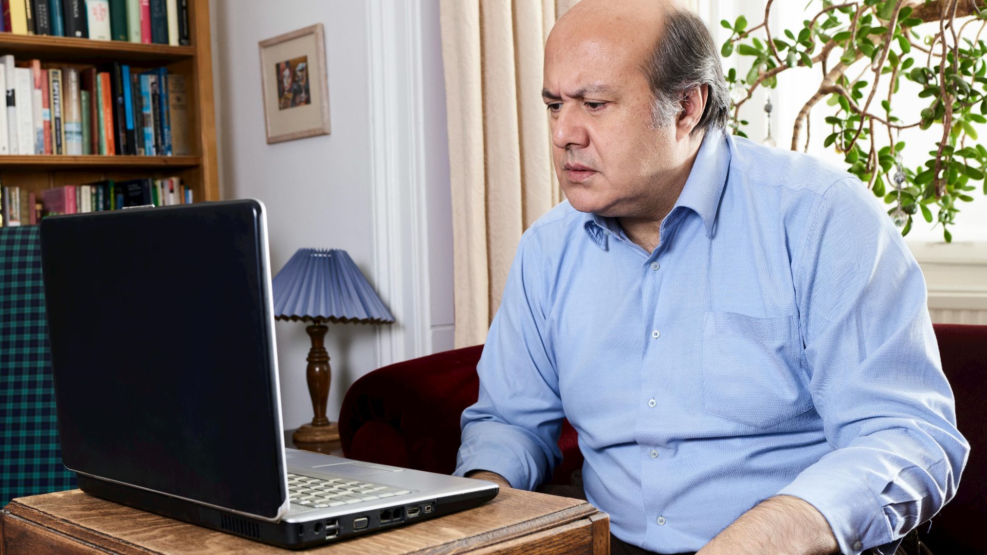 Middle-aged man focused on laptop in a cozy home library setting, surrounded by books.