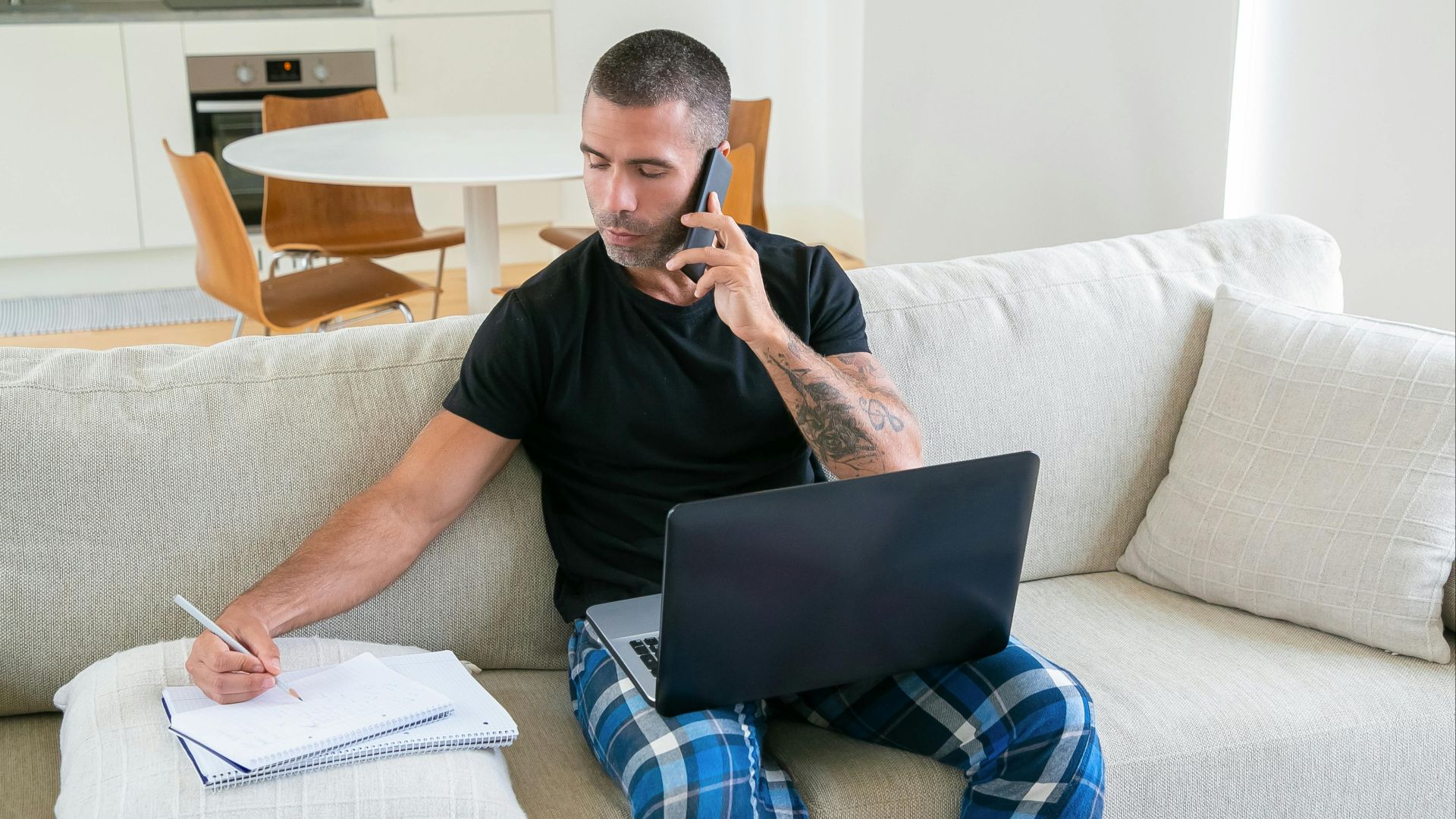 Man multitasking with laptop and smartphone while taking notes on couch at home.