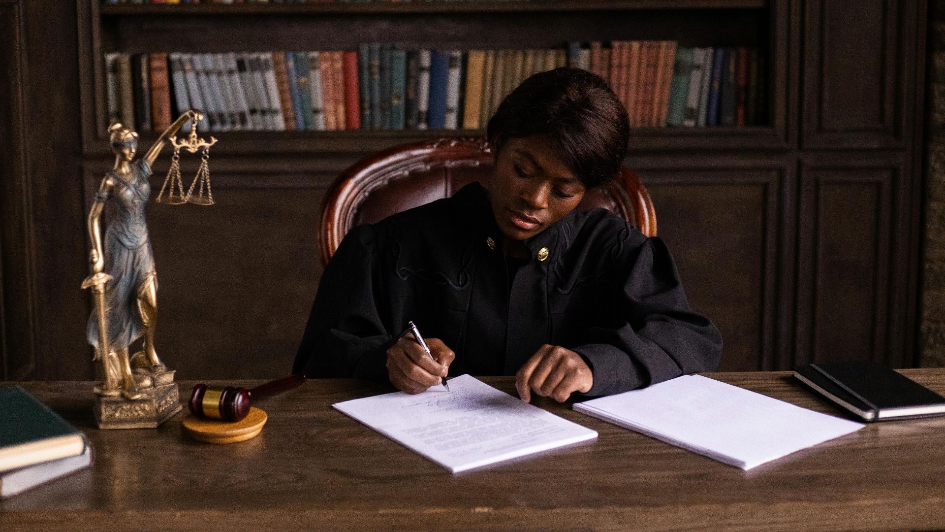 A judge in robes writing on a document at a desk in an office library with law books.