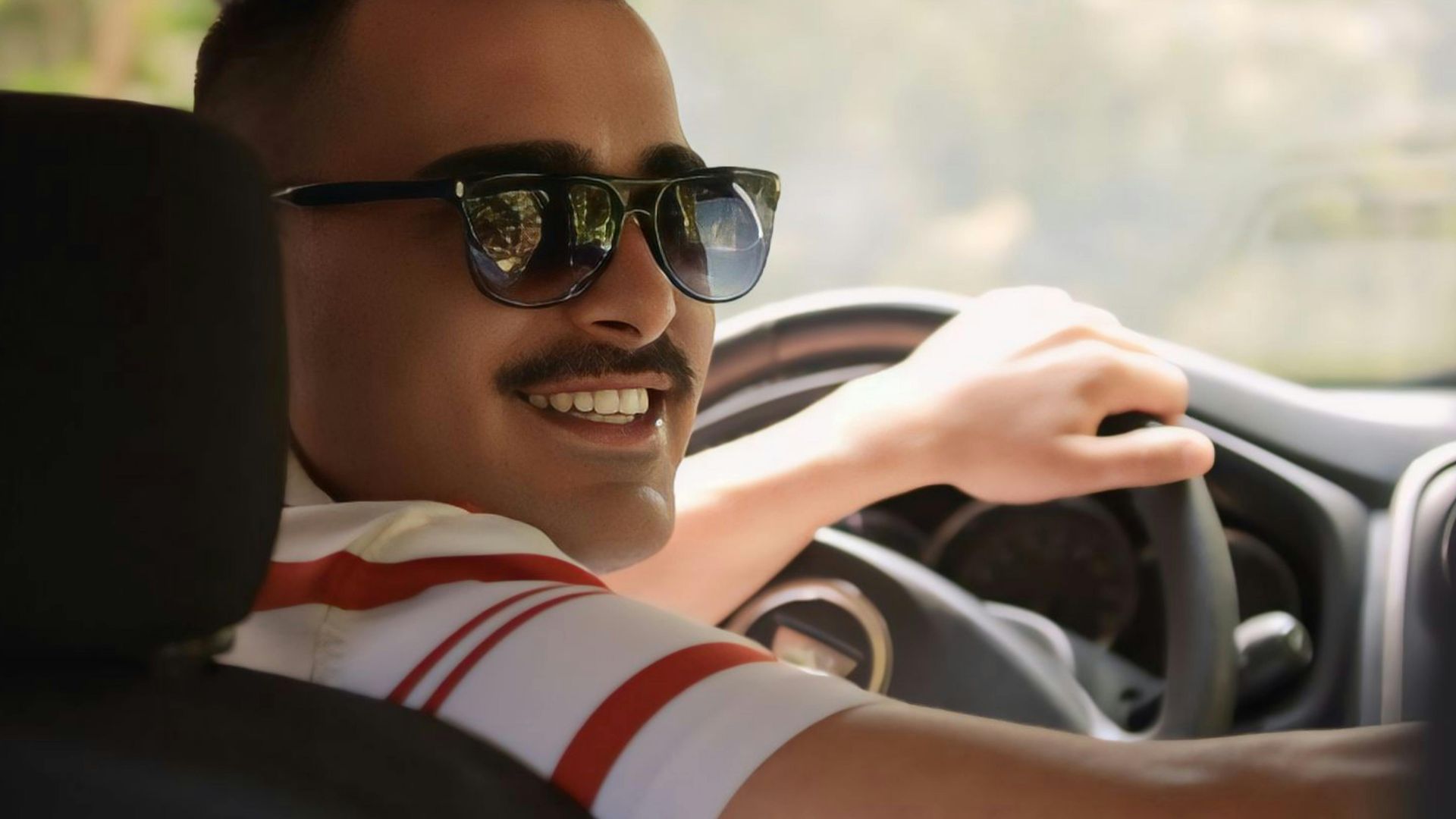 Smiling man wearing sunglasses driving a car with sunlight streaming through the window.