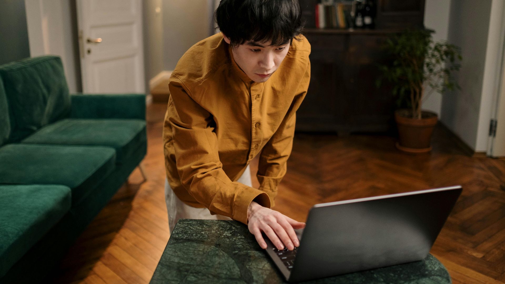 A man standing at a laptop in a stylish living room. Ideal for themes of home office or online learning.