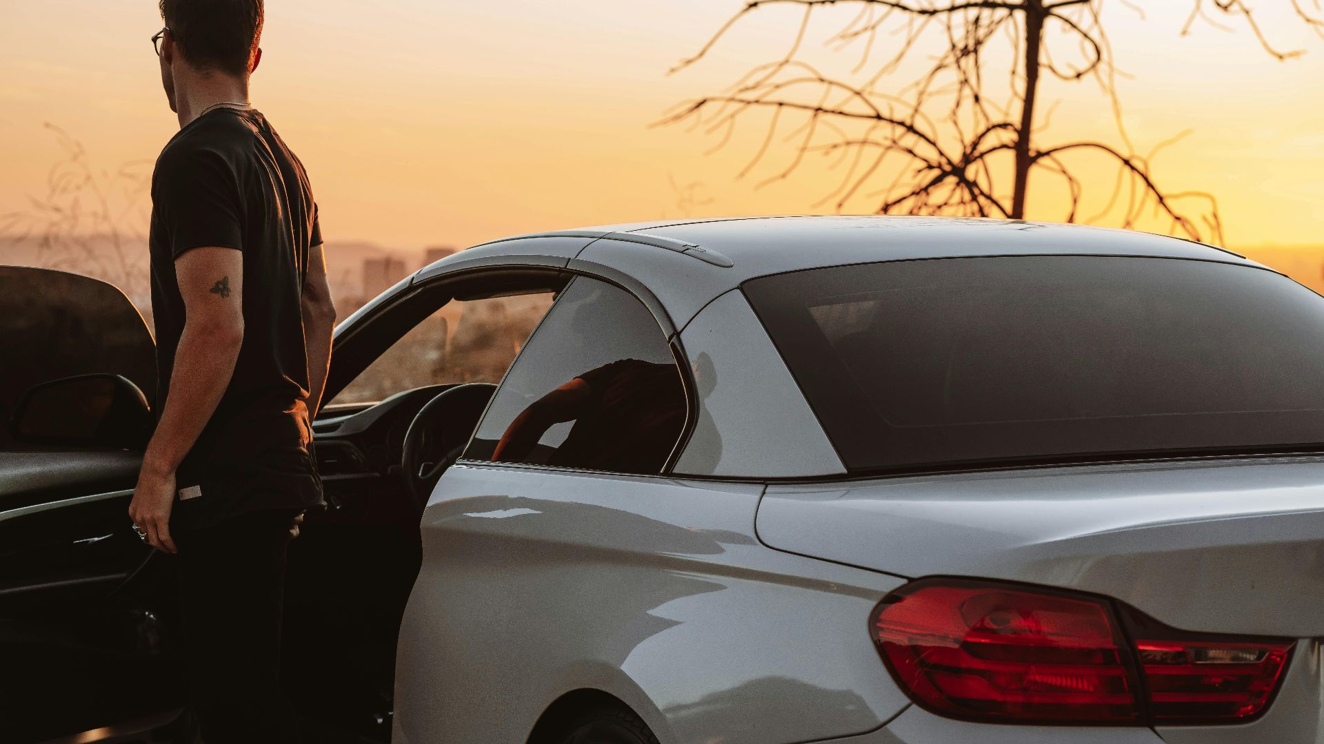 A man stands beside his car, enjoying a scenic sunset in an open landscape.