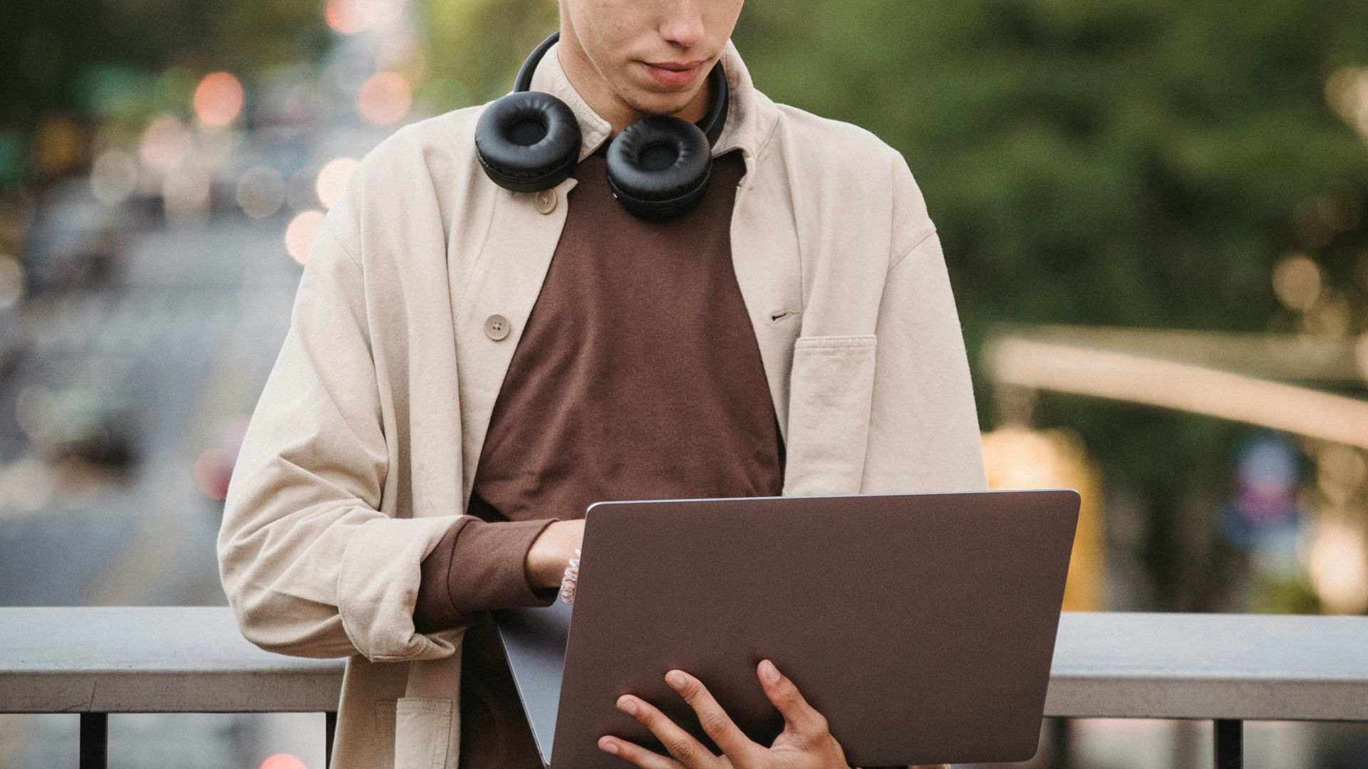 Concentrated young male in casual wear with headphones on neck standing near fence on footbridge over road and using laptop