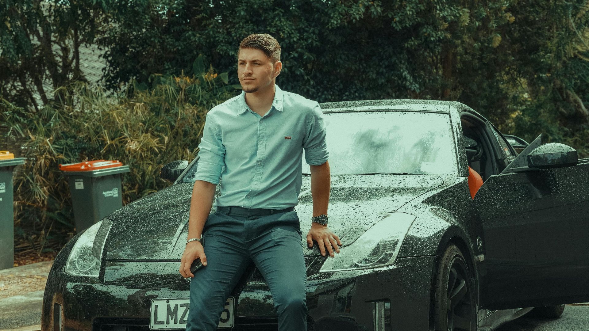 Young man in formal attire leaning on a luxury sports car parked outdoors, exuding style and confidence.