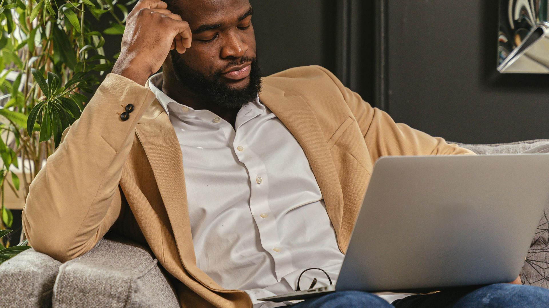 Man working remotely on laptop while relaxing on couch, surrounded by indoor plants.