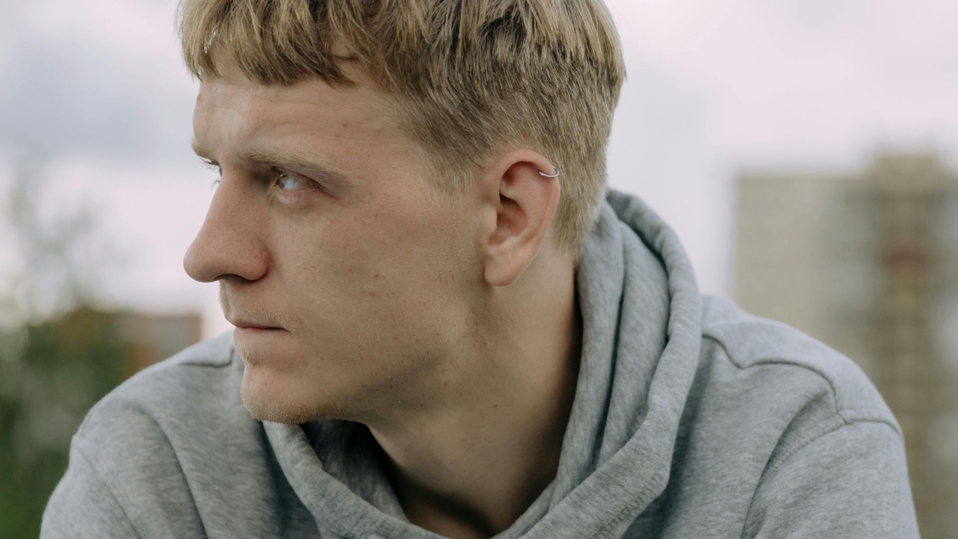 A young man in a gray hoodie looking thoughtfully into the distance outdoors.