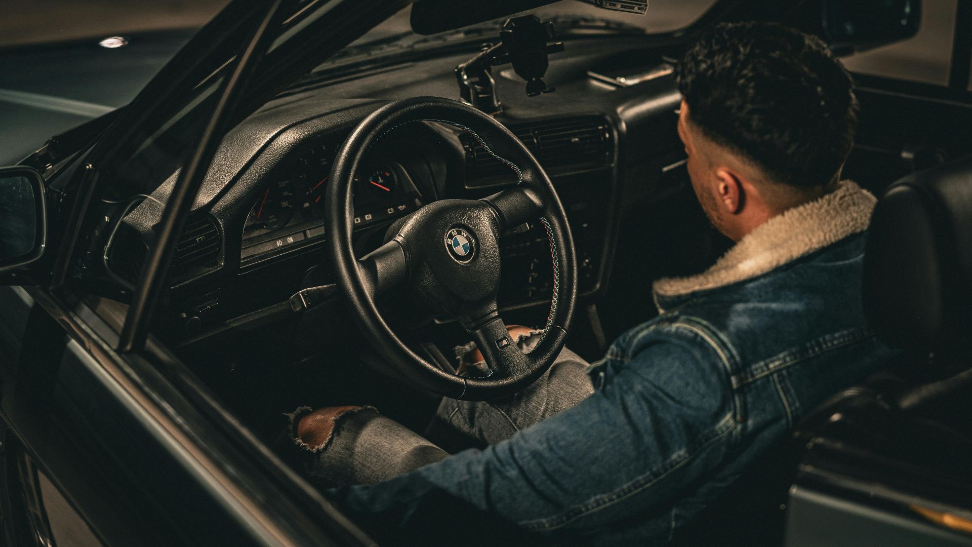 A man in a denim jacket driving an open-top convertible at night. Cozy atmosphere with dim lighting.