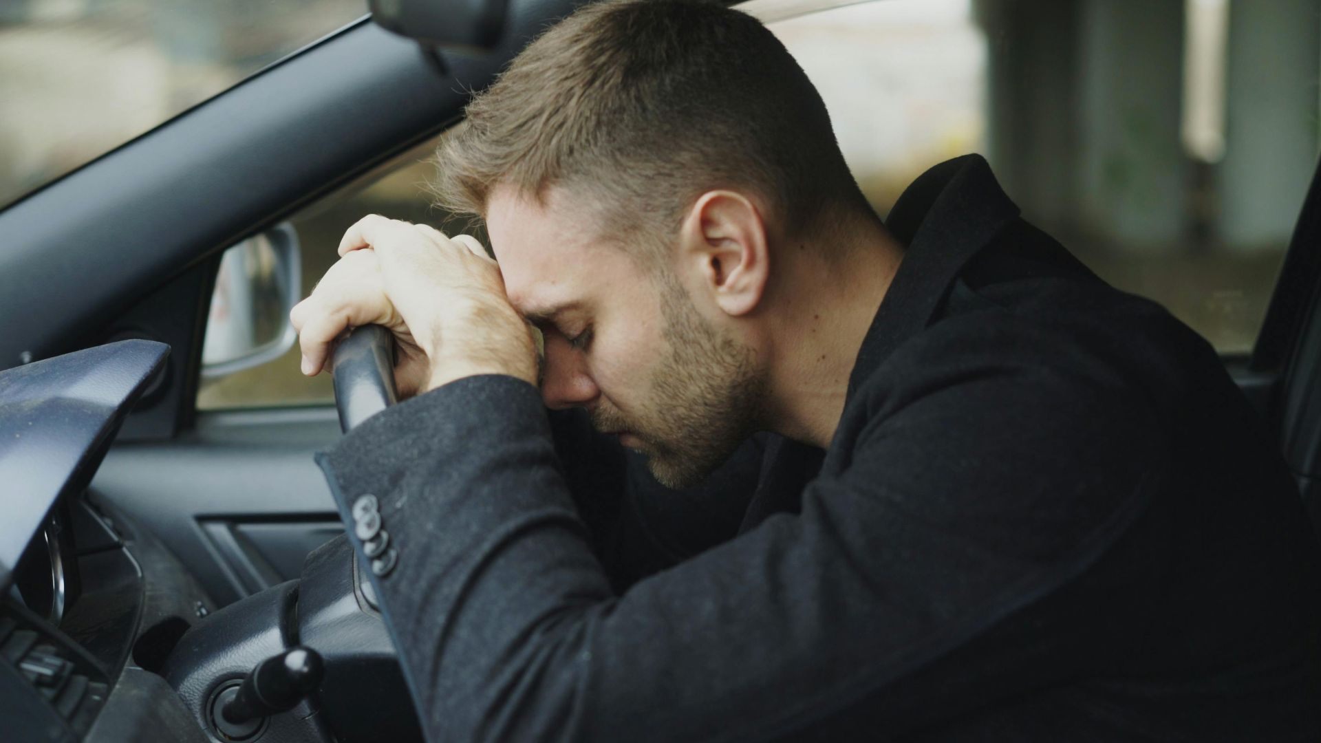A man showing stress while sitting in a car, head on steering wheel.