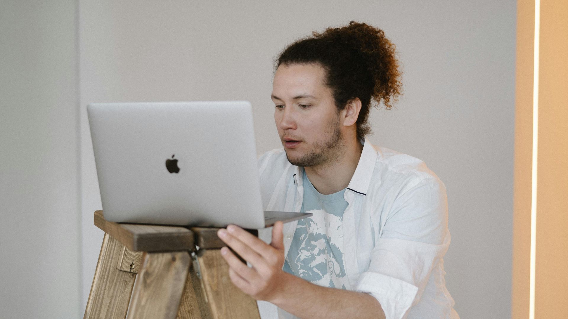 A man using a laptop while standing on a wooden ladder in a minimal indoor setting.