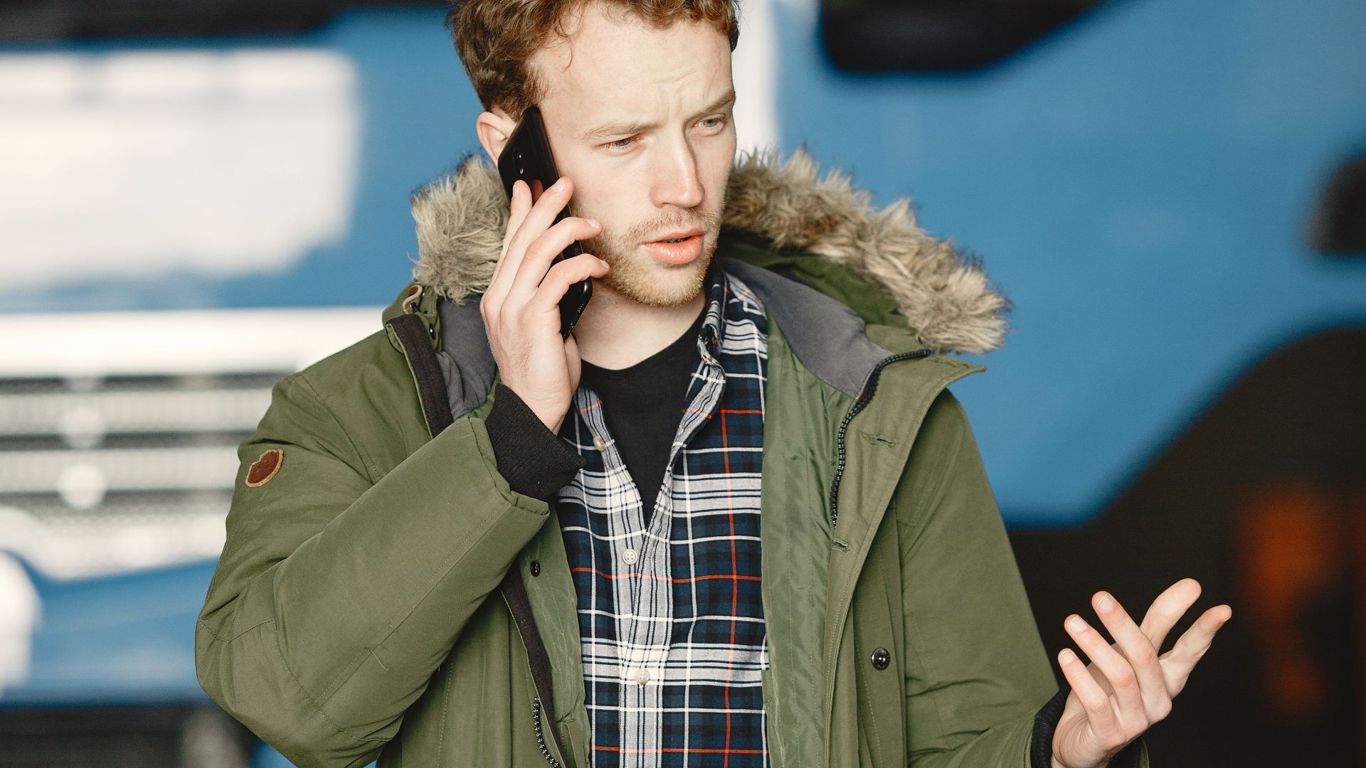 Man wearing green jacket and plaid shirt talks on the phone near a truck outdoors.