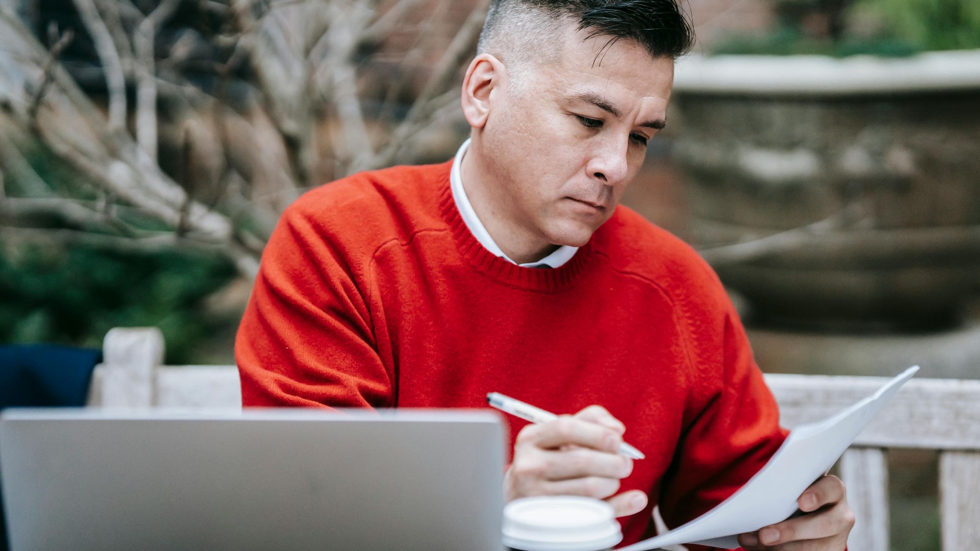 A man in a red sweater concentrating on paperwork while working remotely with a laptop outdoors.