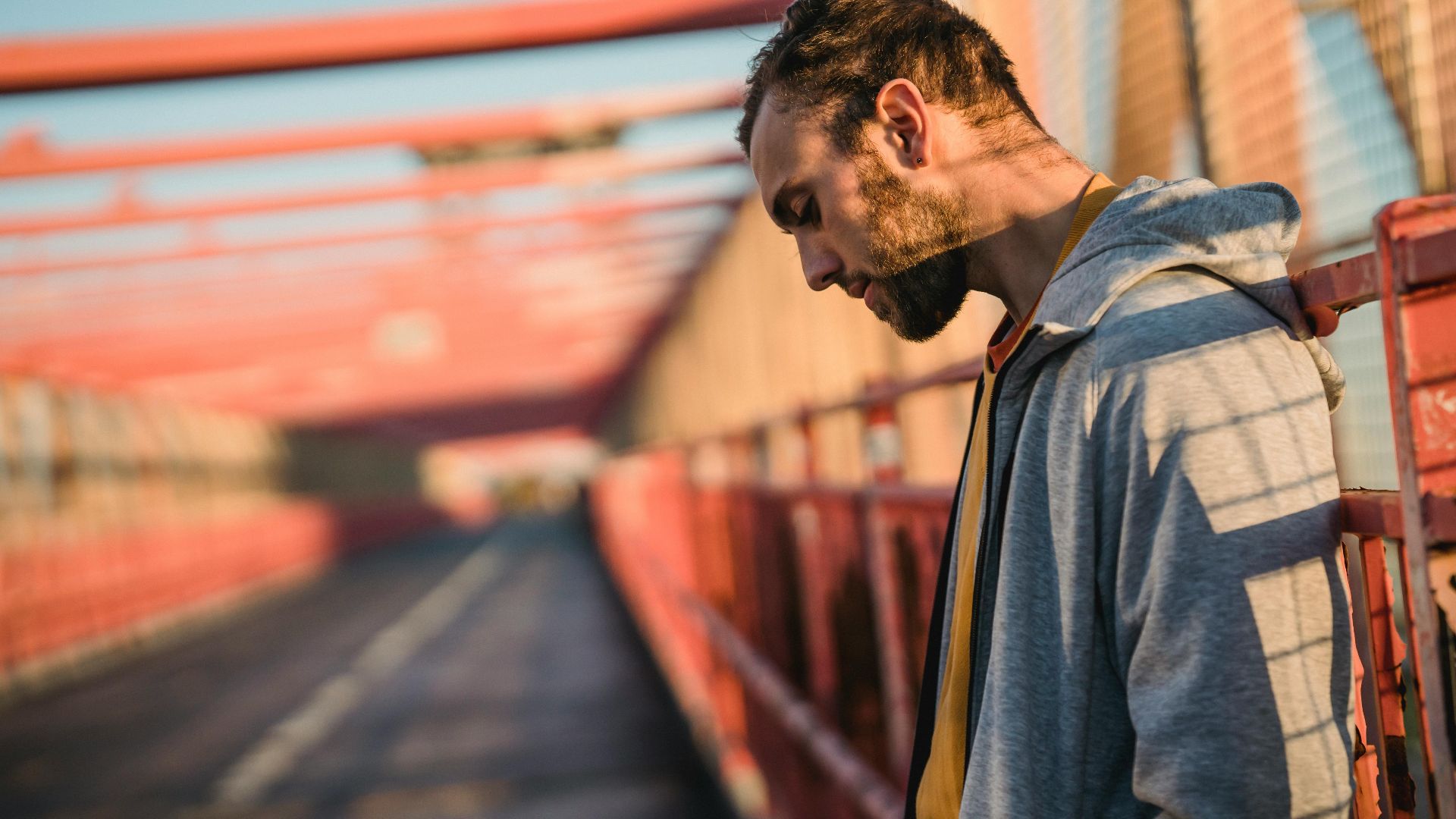 Side view of unshaven millennial guy in hoodie looking down on road of city bridge in sunlight
