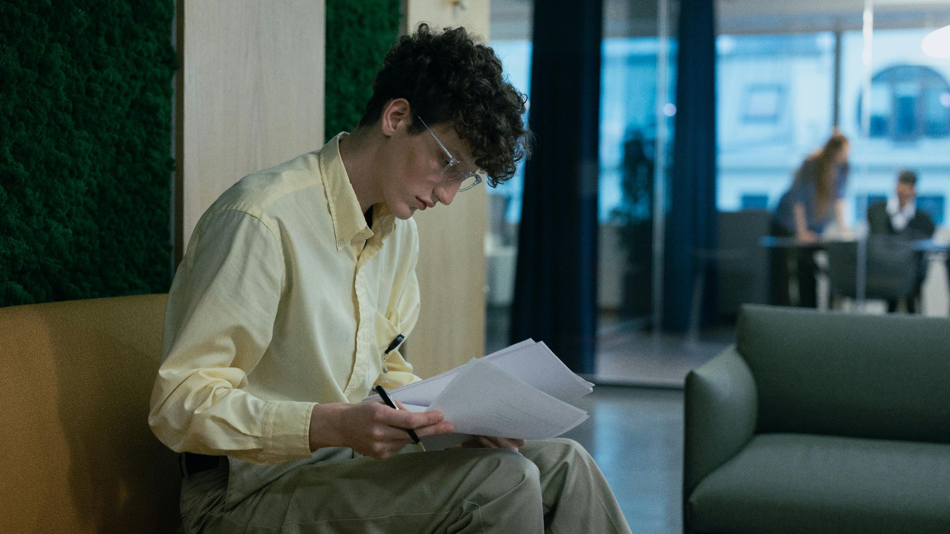 A focused young professional reading documents on a couch in a modern office environment.