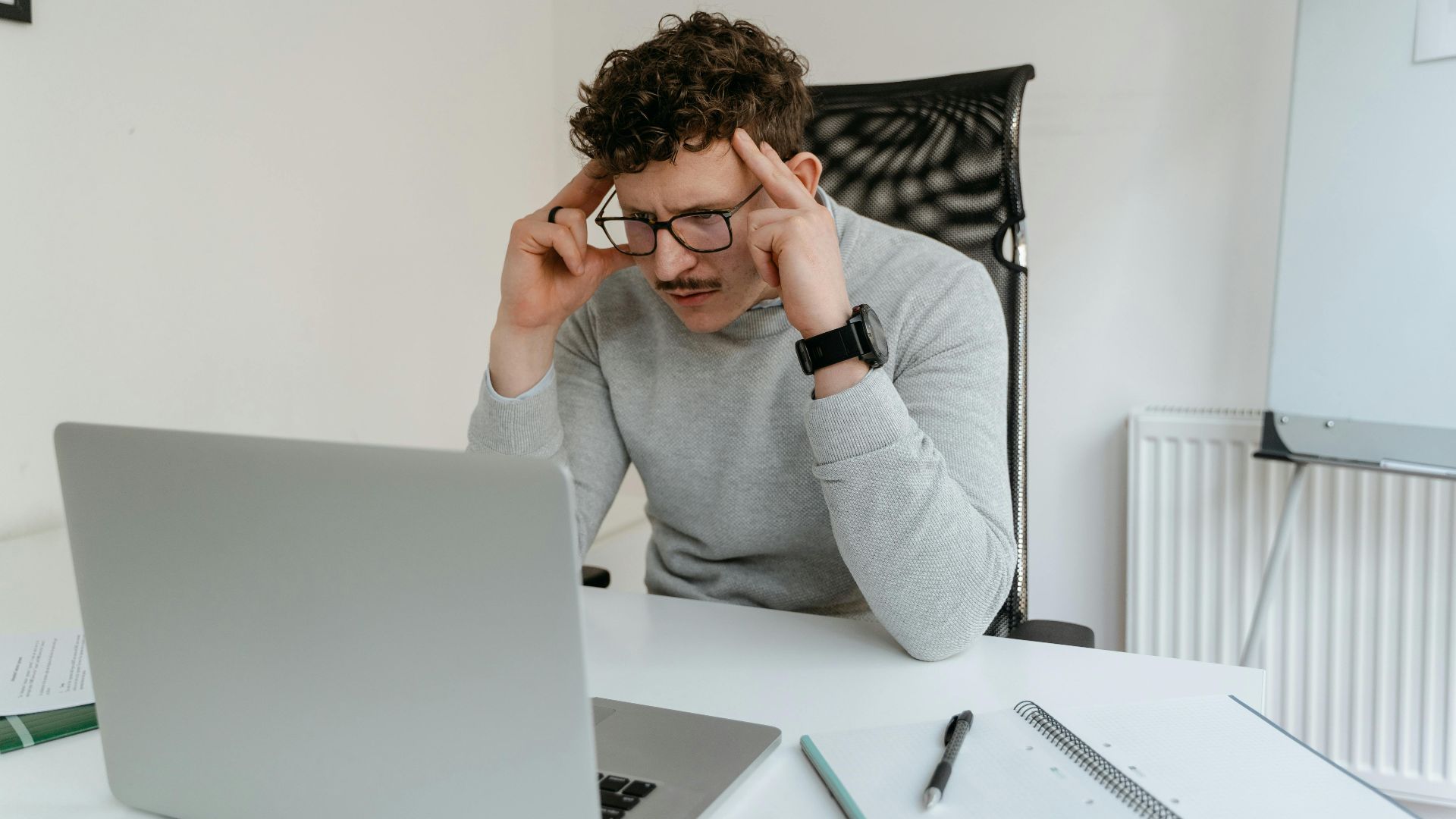 Curly-haired man concentrates while working on a laptop in a modern office setting.