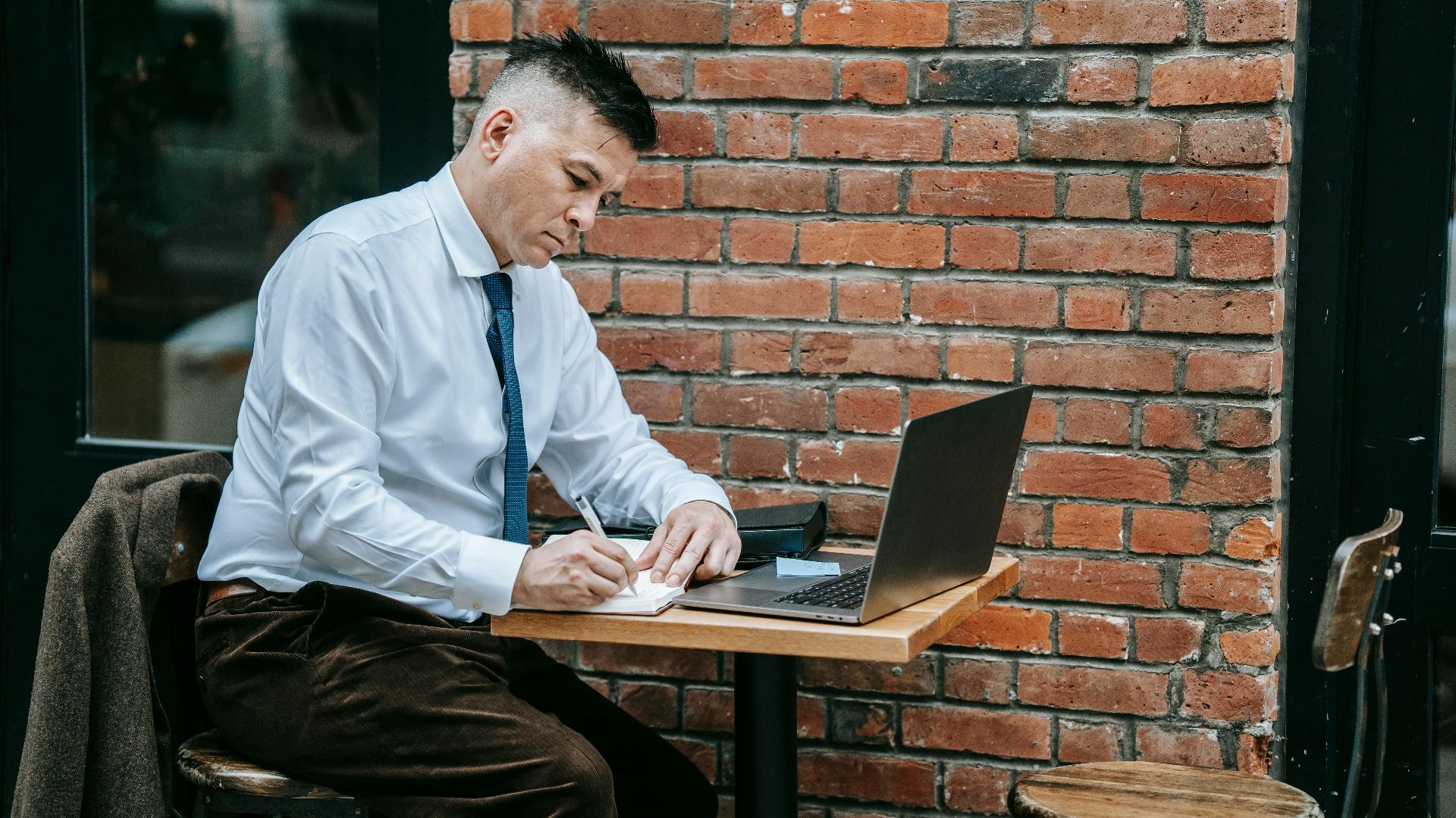 Adult man in corporate attire working on laptop at a cafe table outdoors.